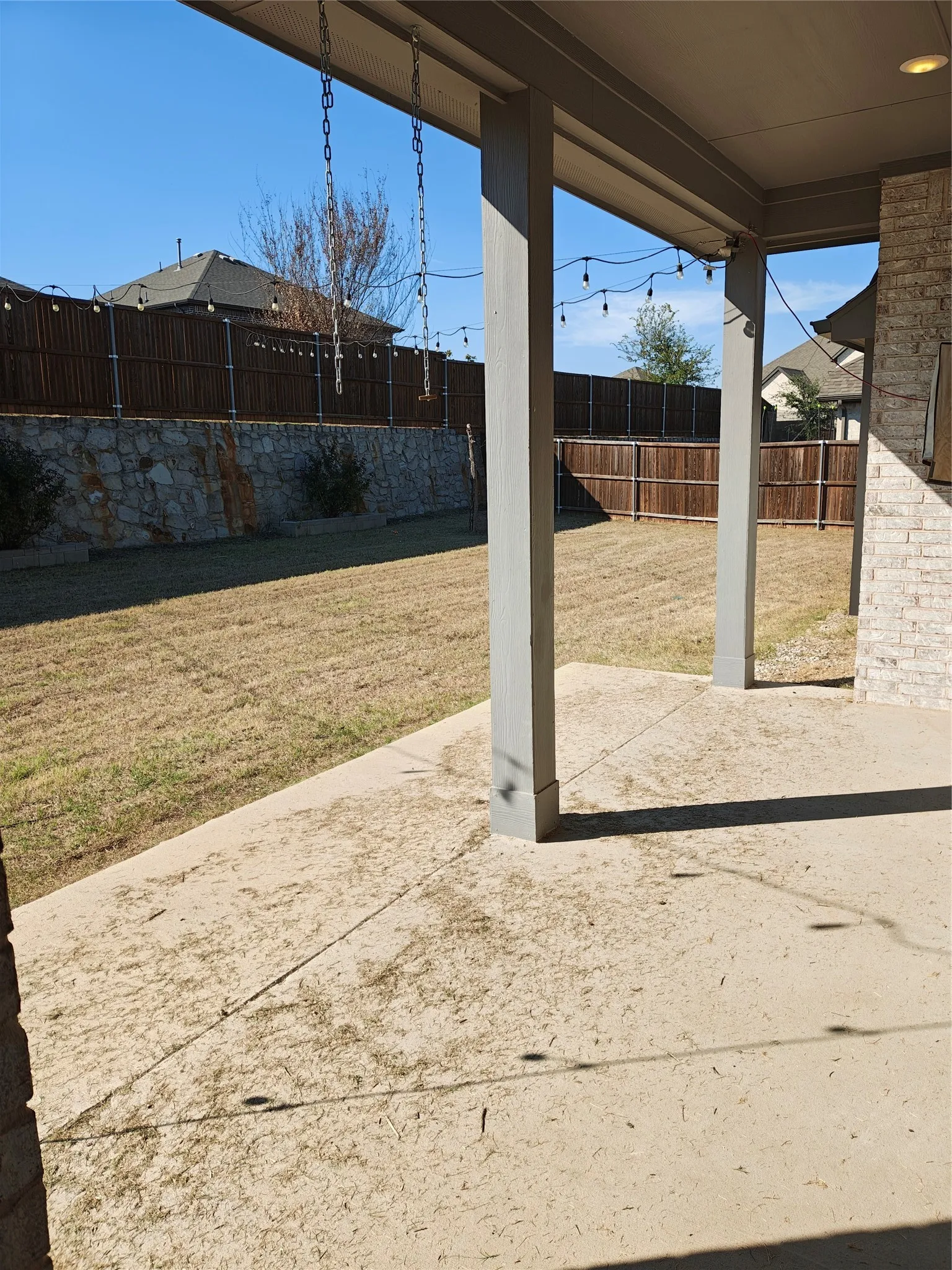 Covered patio with concrete flooring, supported by square columns, and a large grassy yard featuring a stone retaining wall and wood fencing