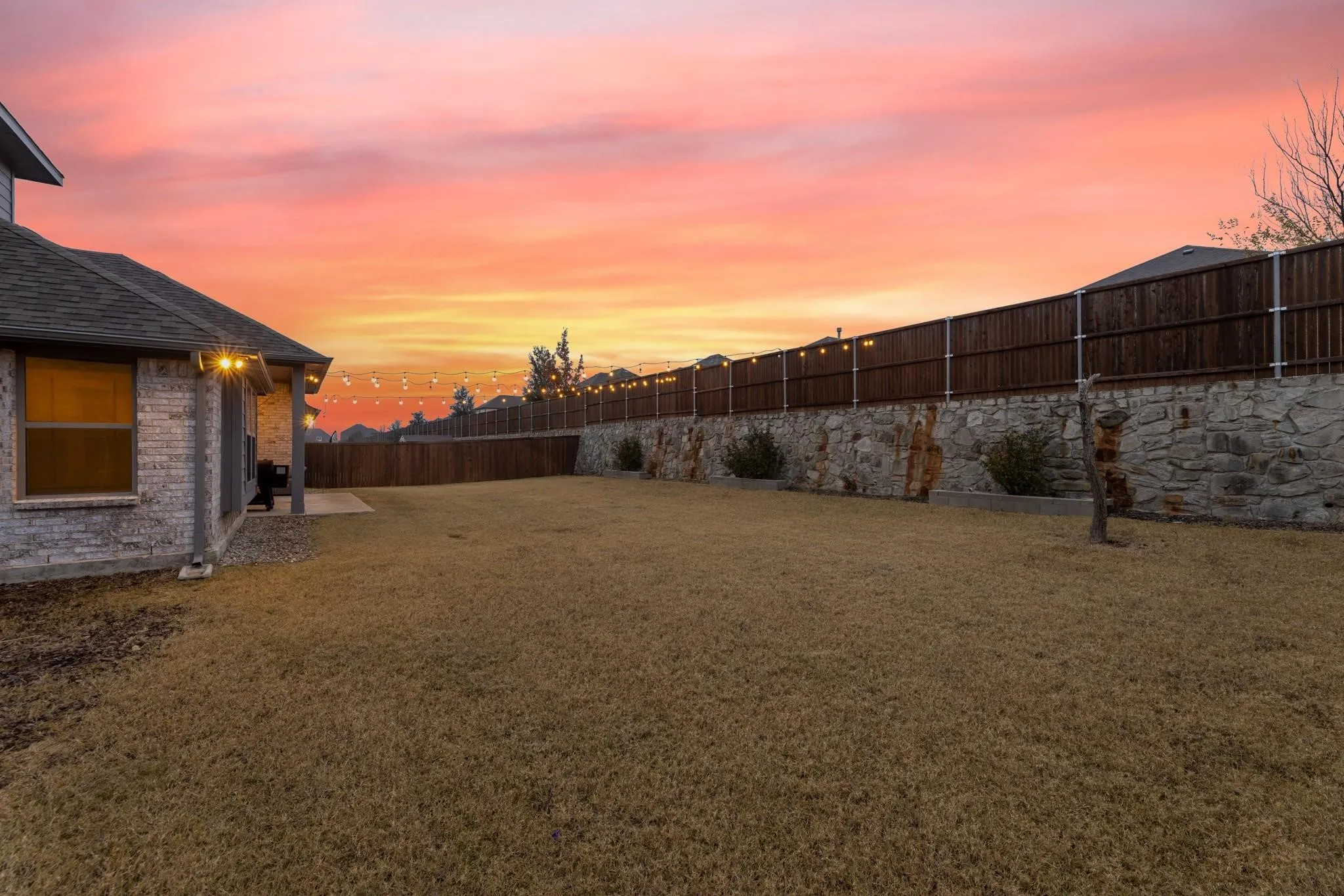 Expansive backyard featuring a stone retaining wall, wood privacy fence, and string lighting
