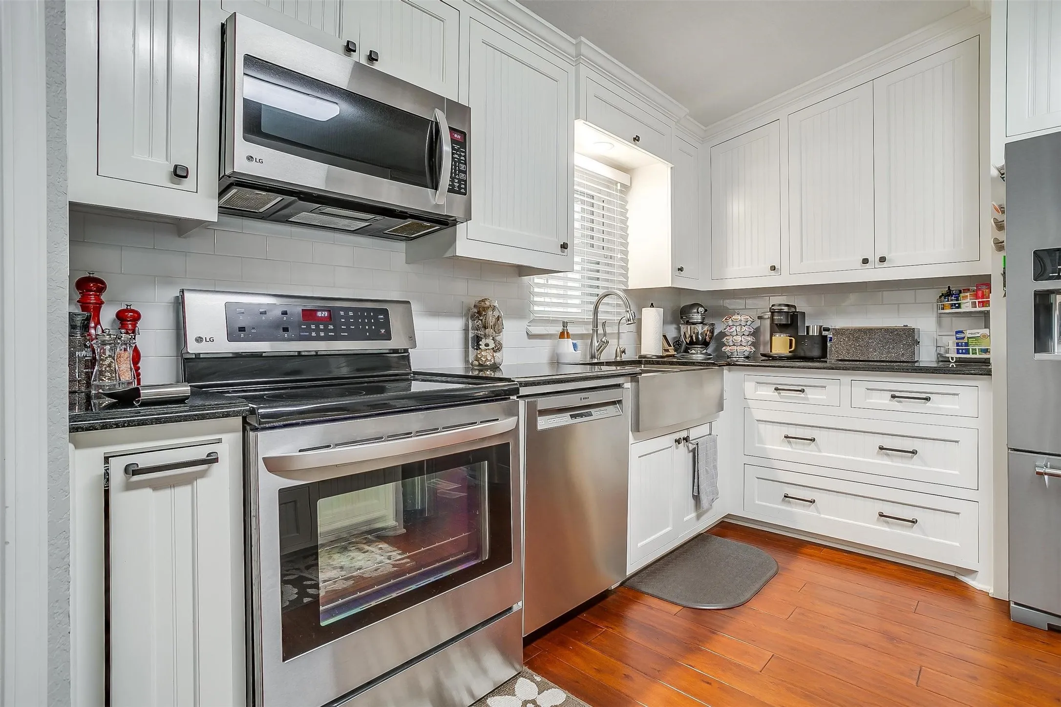 Kitchen featuring white shaker-style cabinetry, dark countertops, stainless steel appliances, a farmhouse sink, and wood-finish flooring