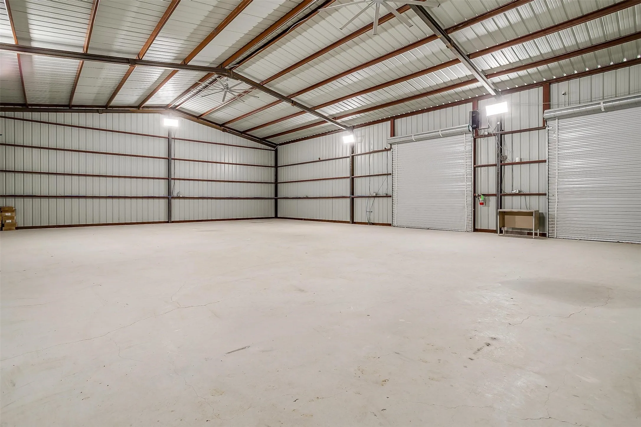 Spacious metal building featuring a concrete floor, exposed steel beam construction, corrugated metal walls, multiple roll-up doors, and overhead lighting fixtures