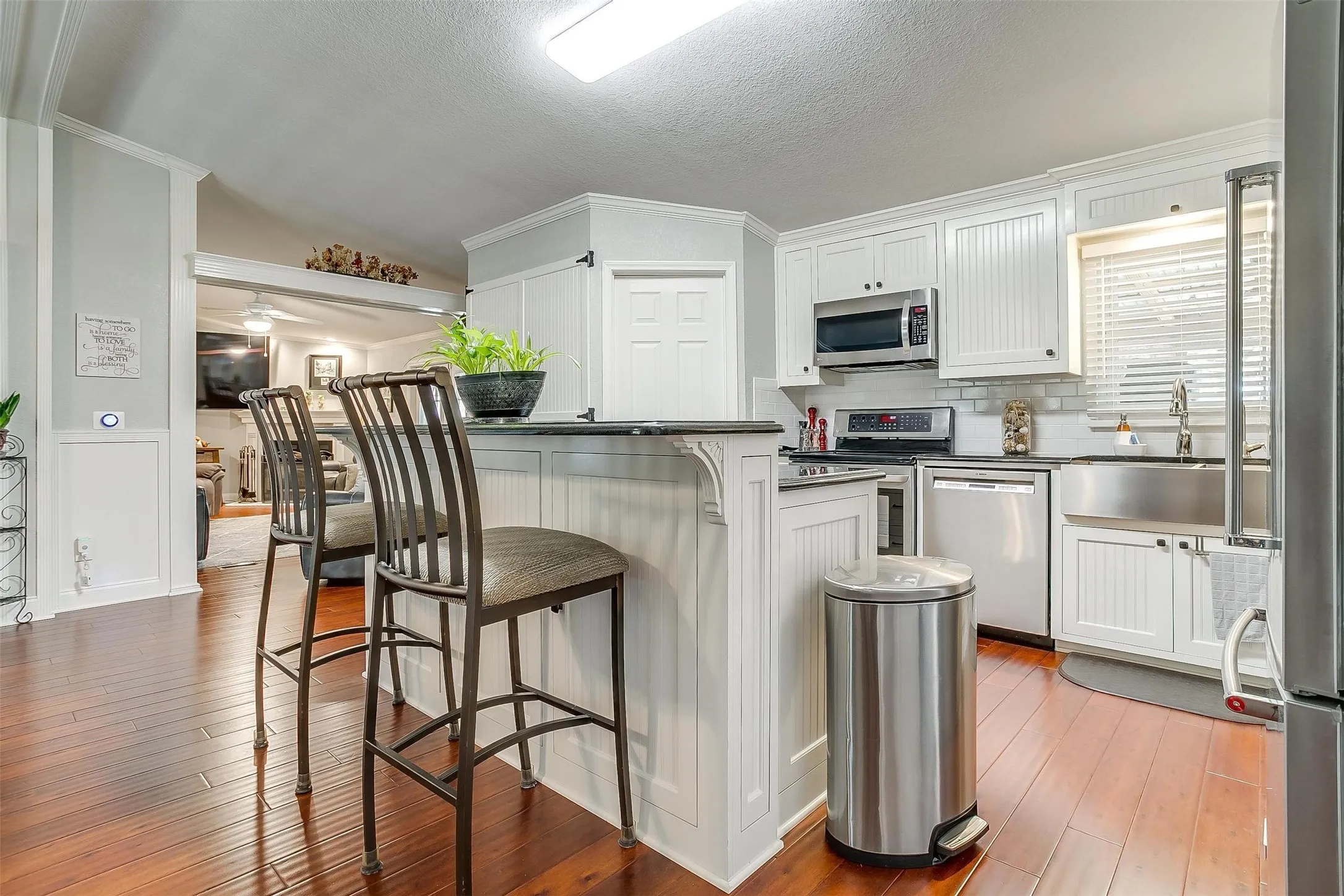 Kitchen featuring rich wood-finish flooring, white beadboard cabinetry, stainless steel appliances, a farmhouse-style sink, and a breakfast bar with dark countertops