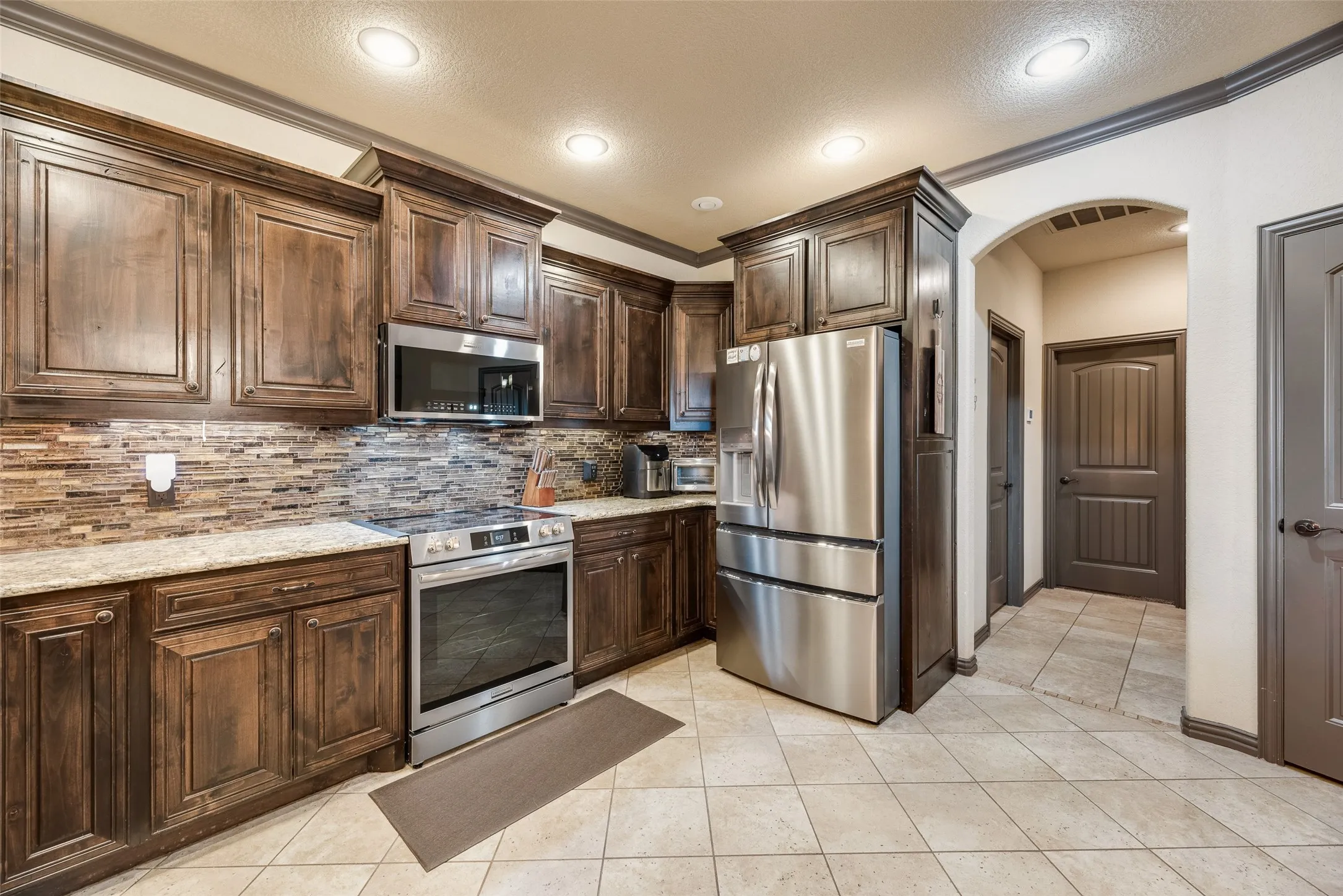 Modern kitchen with stainless steel appliances, rich wood cabinetry, patterned tile backsplash, and light-toned tile flooring