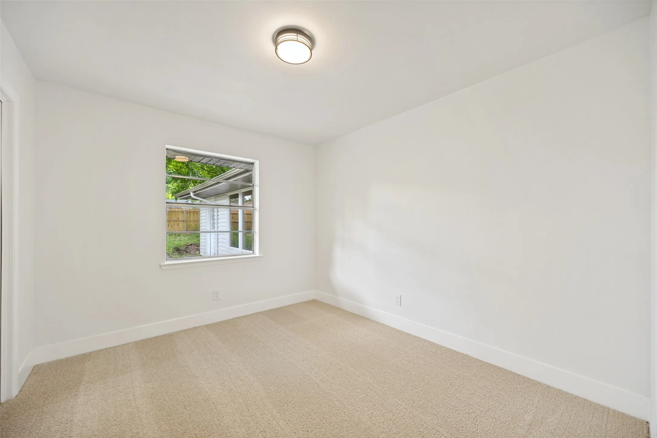 Interior room featuring light-colored carpeting, white baseboards, and a single window with exterior landscaping visible