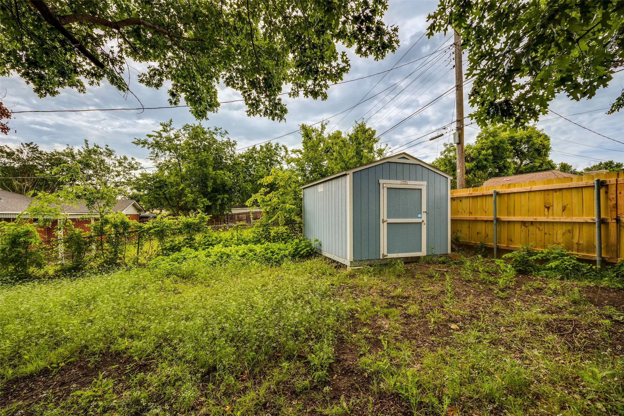Backyard featuring a light blue storage shed with a white trim door, a wooden privacy fence, and mature trees