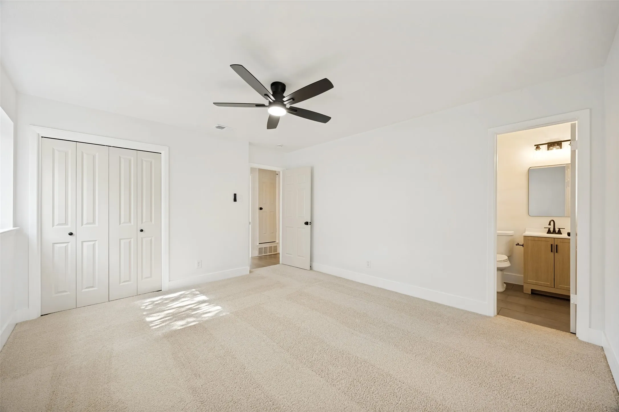 Carpeted bedroom featuring white walls, a black ceiling fan with integrated lighting, bi-fold closet doors, and an en-suite bathroom entrance