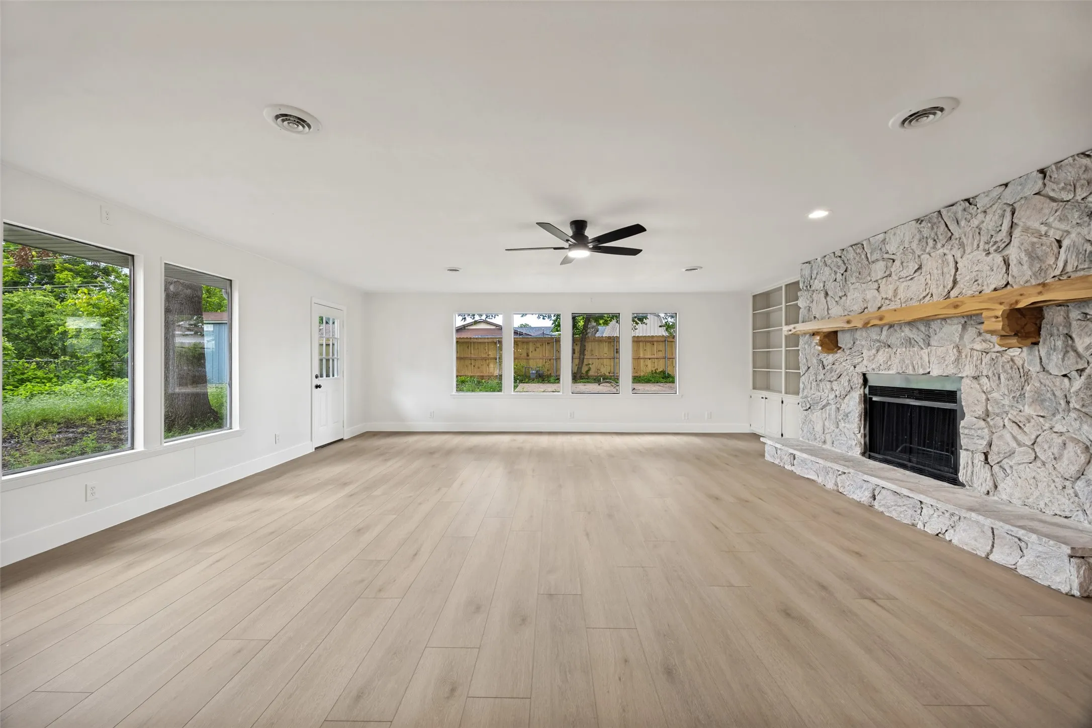 Spacious room featuring wood-finish flooring, a stone fireplace with a substantial timber mantel, built-in shelving, a ceiling fan, and multiple windows