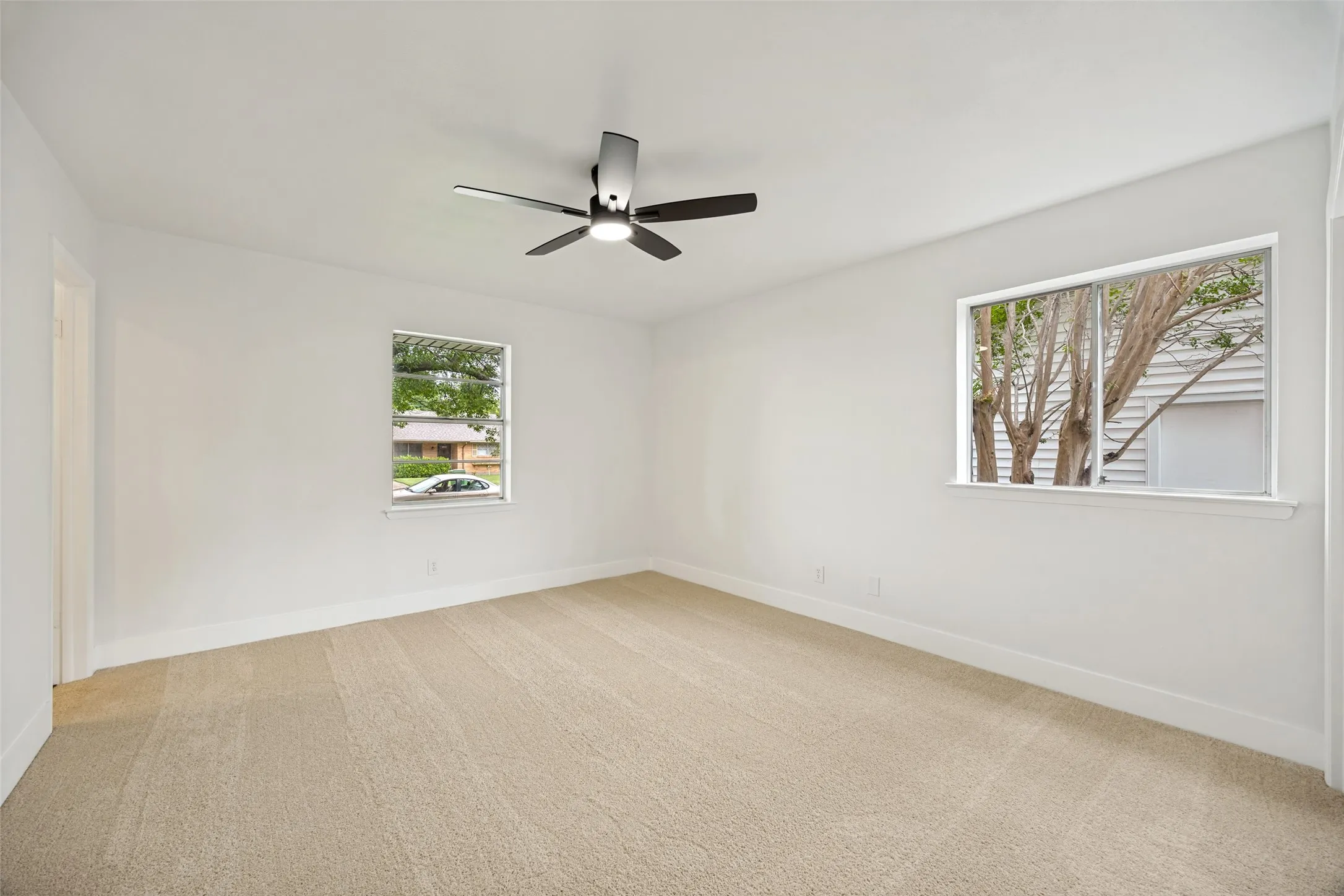 Spacious room featuring neutral carpeting, white walls, two windows, and a modern ceiling fan with integrated lighting