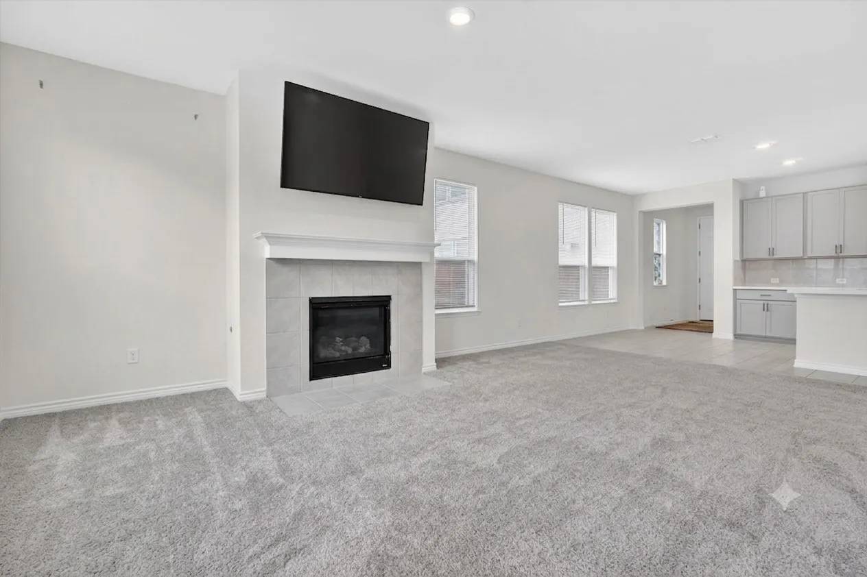 Spacious living area featuring a fireplace with a light-toned tile surround and mantel, soft grey carpeting, multiple windows, recessed lighting, and a partial view of the kitchen with light grey cabinetry