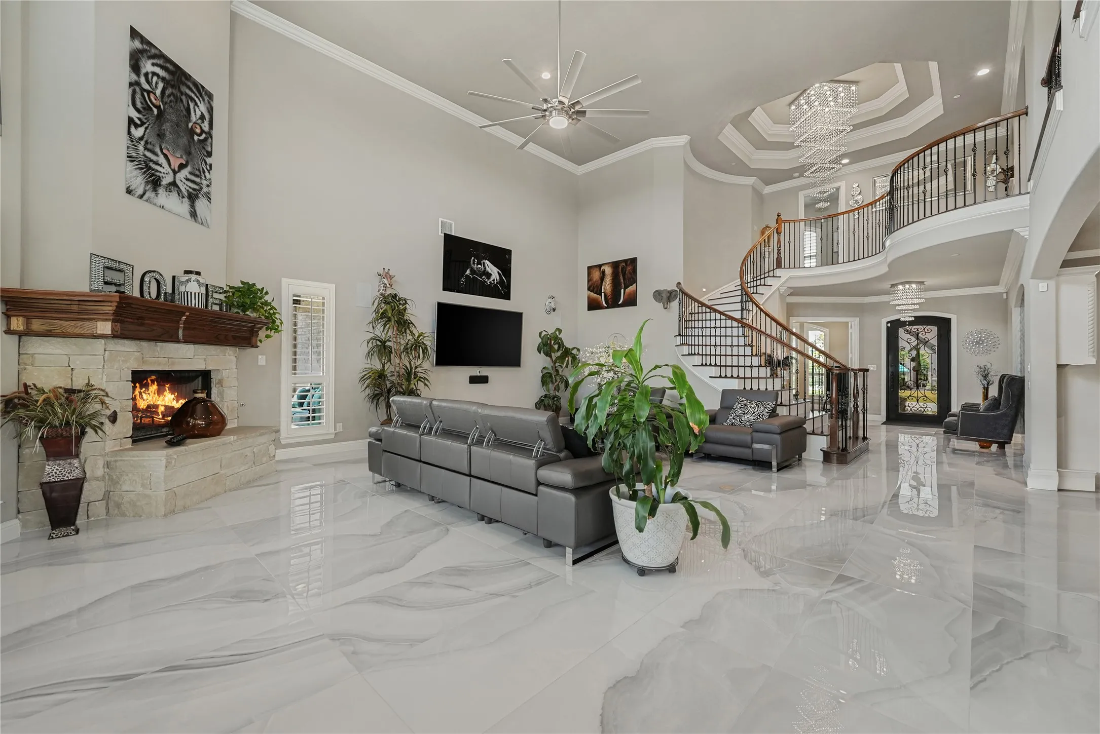 Spacious living area featuring high vaulted ceilings, large format tile flooring, a stone fireplace with a wood-beam mantel, a spiral staircase with wrought iron balusters, and a tiered tray ceiling with a chandelier