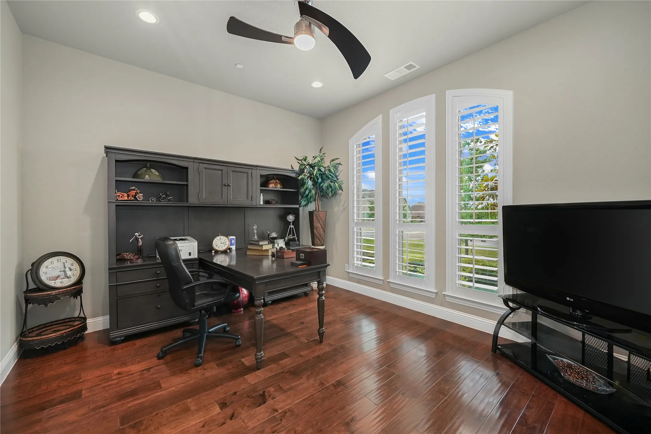 Room featuring warm wood-finish flooring, three arched windows with plantation shutters, a ceiling fan with integrated lighting, and recessed ceiling lights