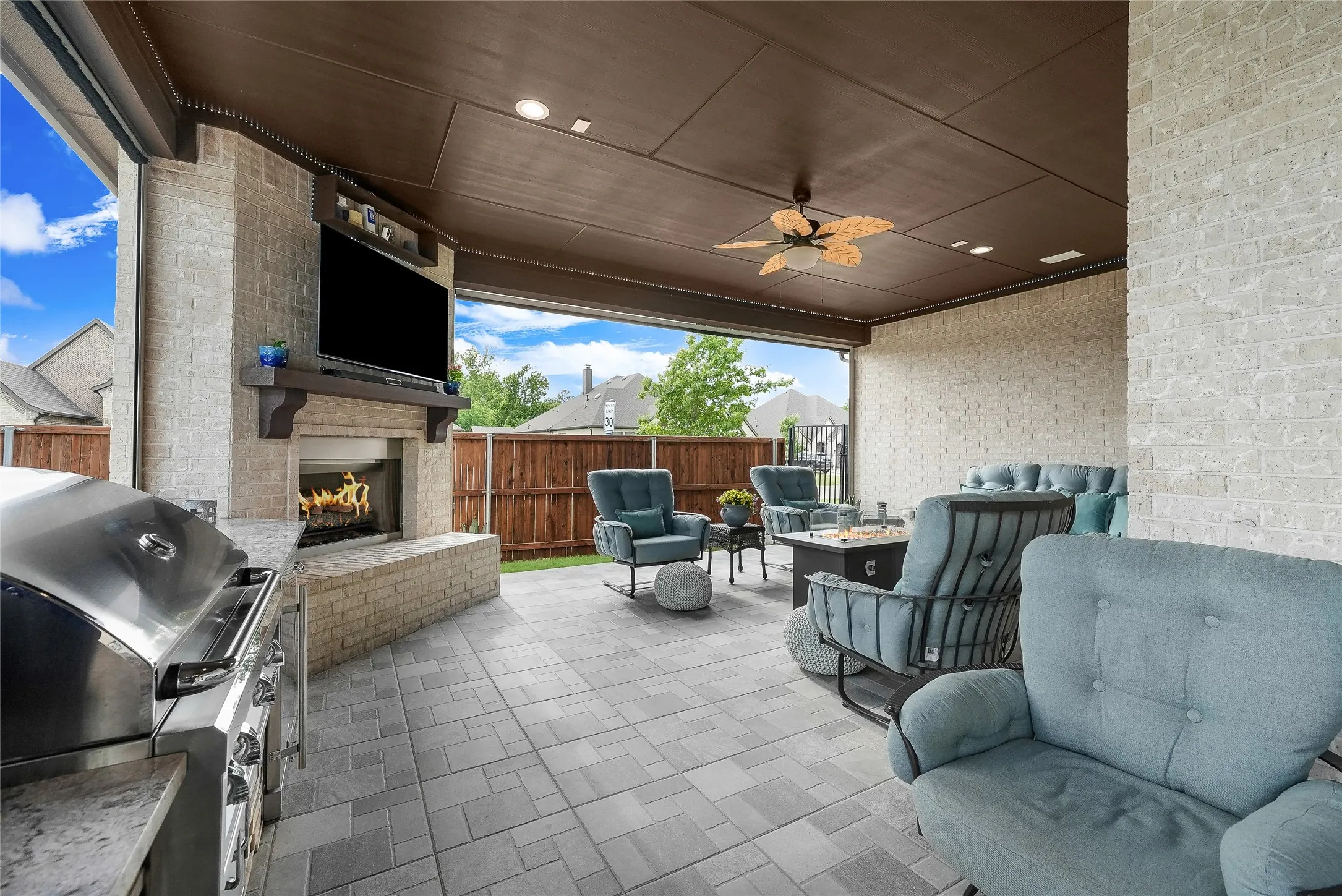 Covered outdoor living space featuring a built-in outdoor kitchen with stainless steel grill, stone fireplace with mantel, ceiling fan, and recessed lighting