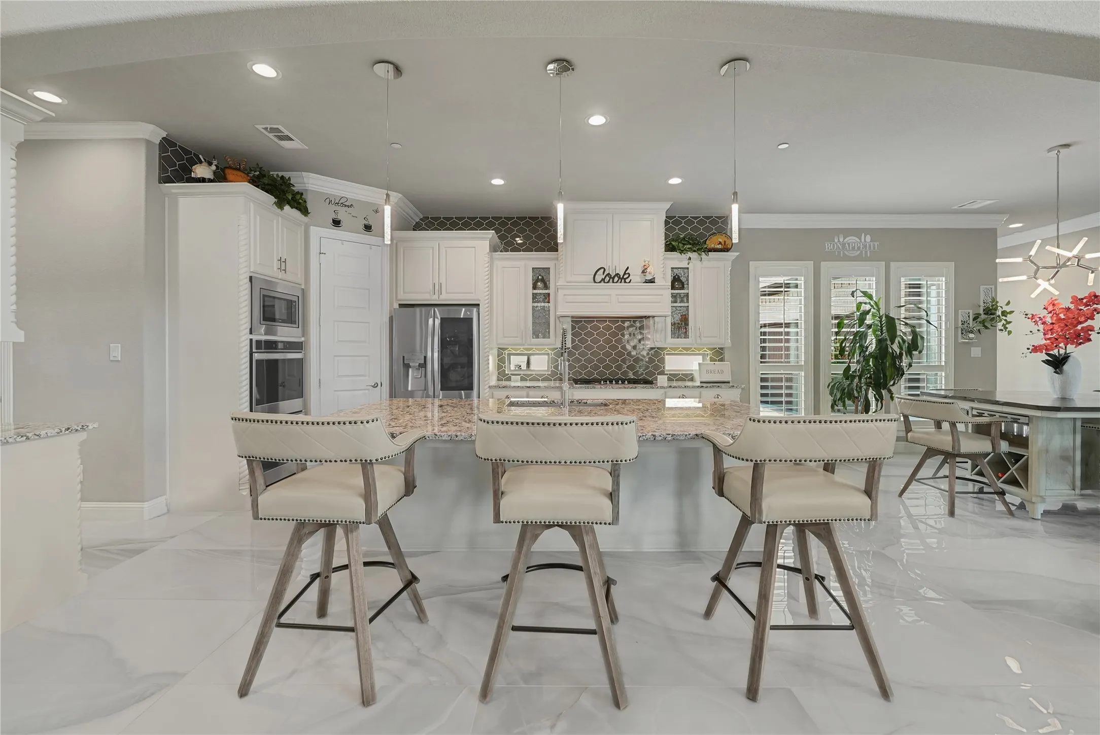 Open-concept kitchen featuring an expansive island with a light-toned countertop, white cabinetry with crown molding, stainless steel built-in appliances, and a decorative backsplash