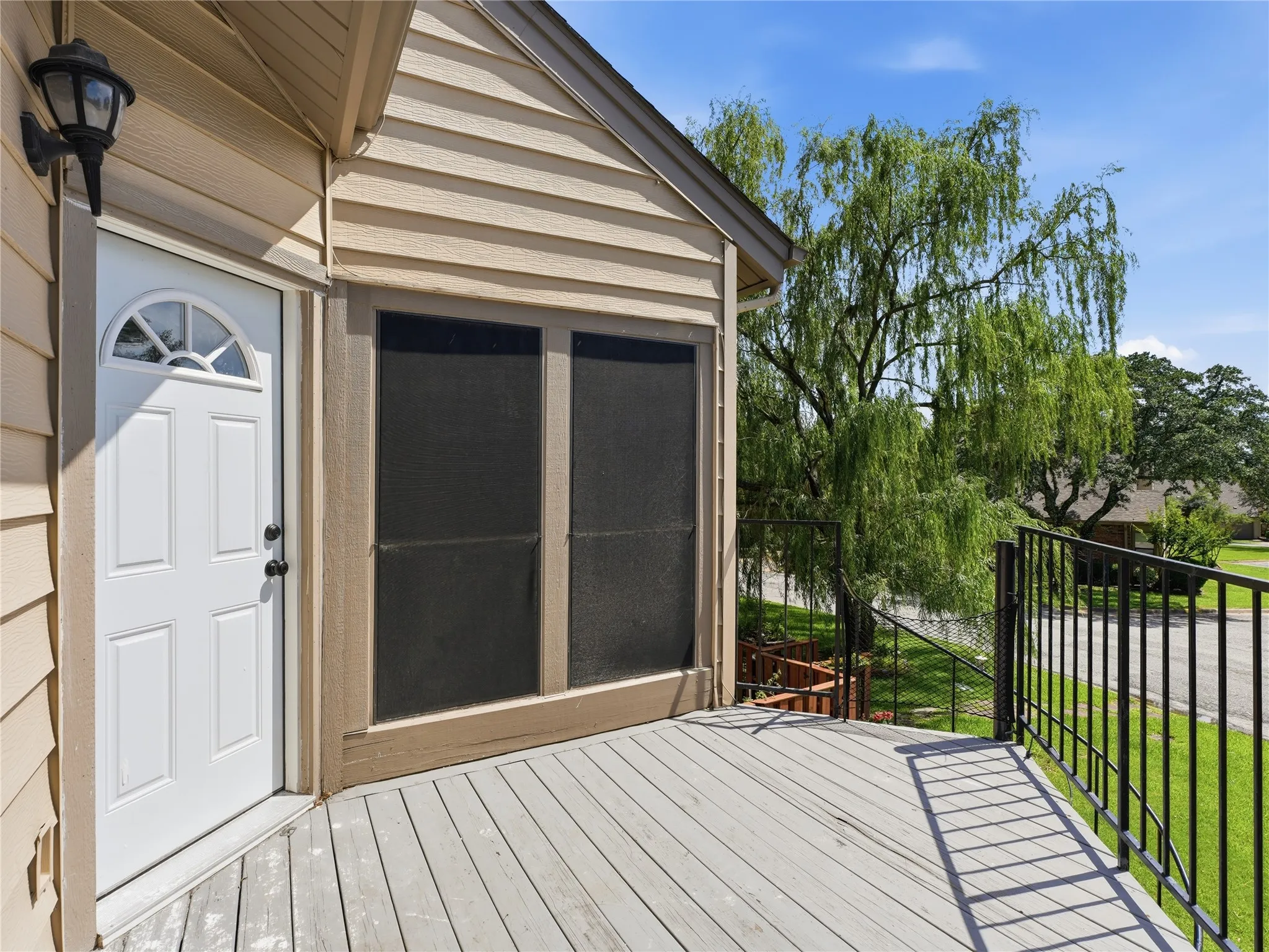 Front porch with weathered wood-finish decking, white paneled entry door with an arched window, decorative exterior light fixture, and black metal railings