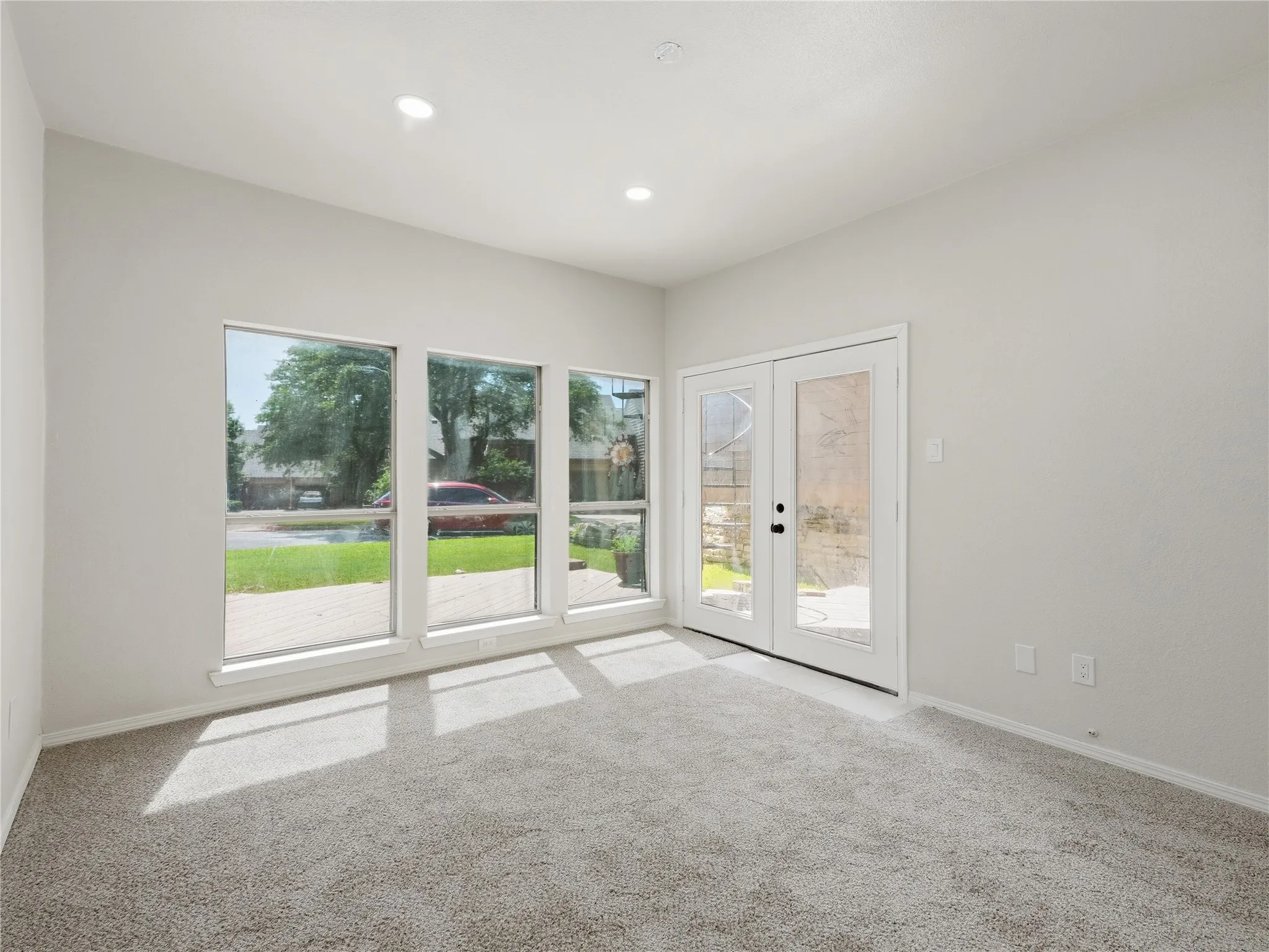 Light gray carpeting, recessed ceiling lighting, multiple large windows, and a set of glass-paneled French doors
