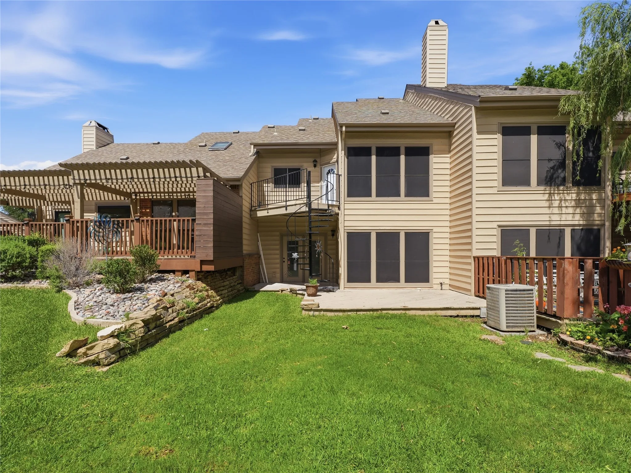 Rear exterior featuring a spacious wood deck with a pergola, a spiraling metal staircase, a lower concrete patio, and a sloped lawn with a rock retaining wall