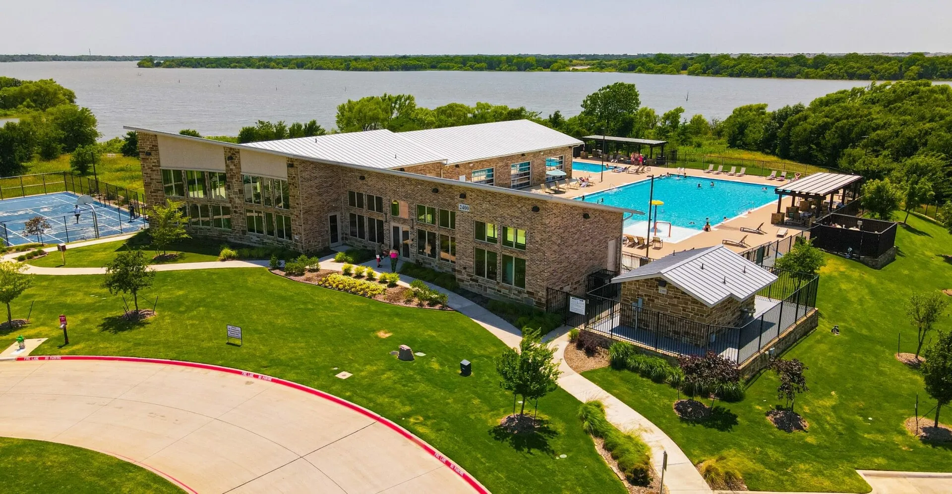Modern stone and brick-clad building featuring extensive glazing, a large outdoor swimming pool, and an adjacent tennis court