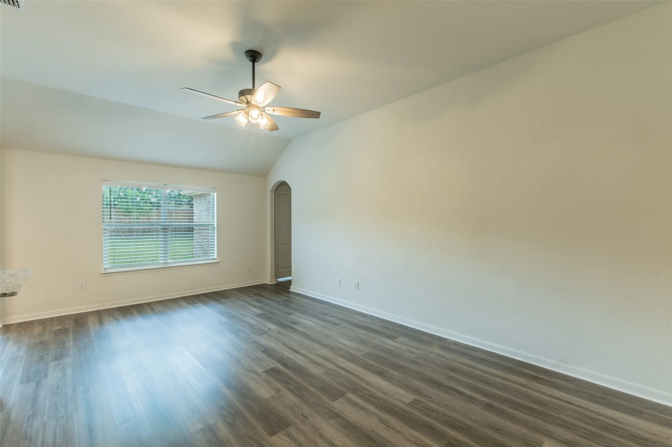 Spacious living area featuring vaulted ceilings, wood-finish flooring, a ceiling fan with integrated lighting, an arched doorway, and a window with blinds