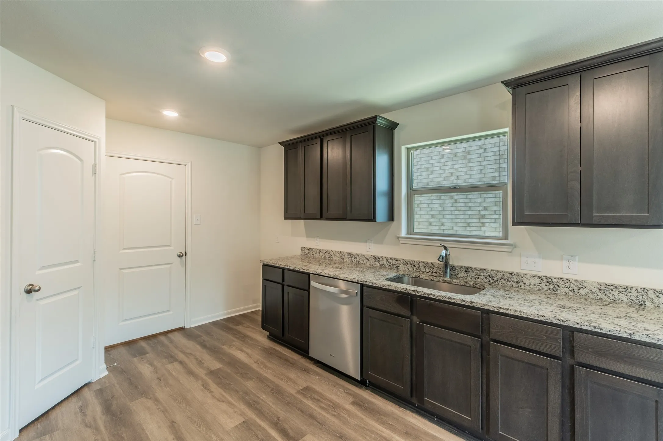 Kitchen featuring wood-finish flooring, dark wood-finish cabinetry, granite-style countertops, stainless steel sink with a single-handle faucet, and a stainless steel dishwasher