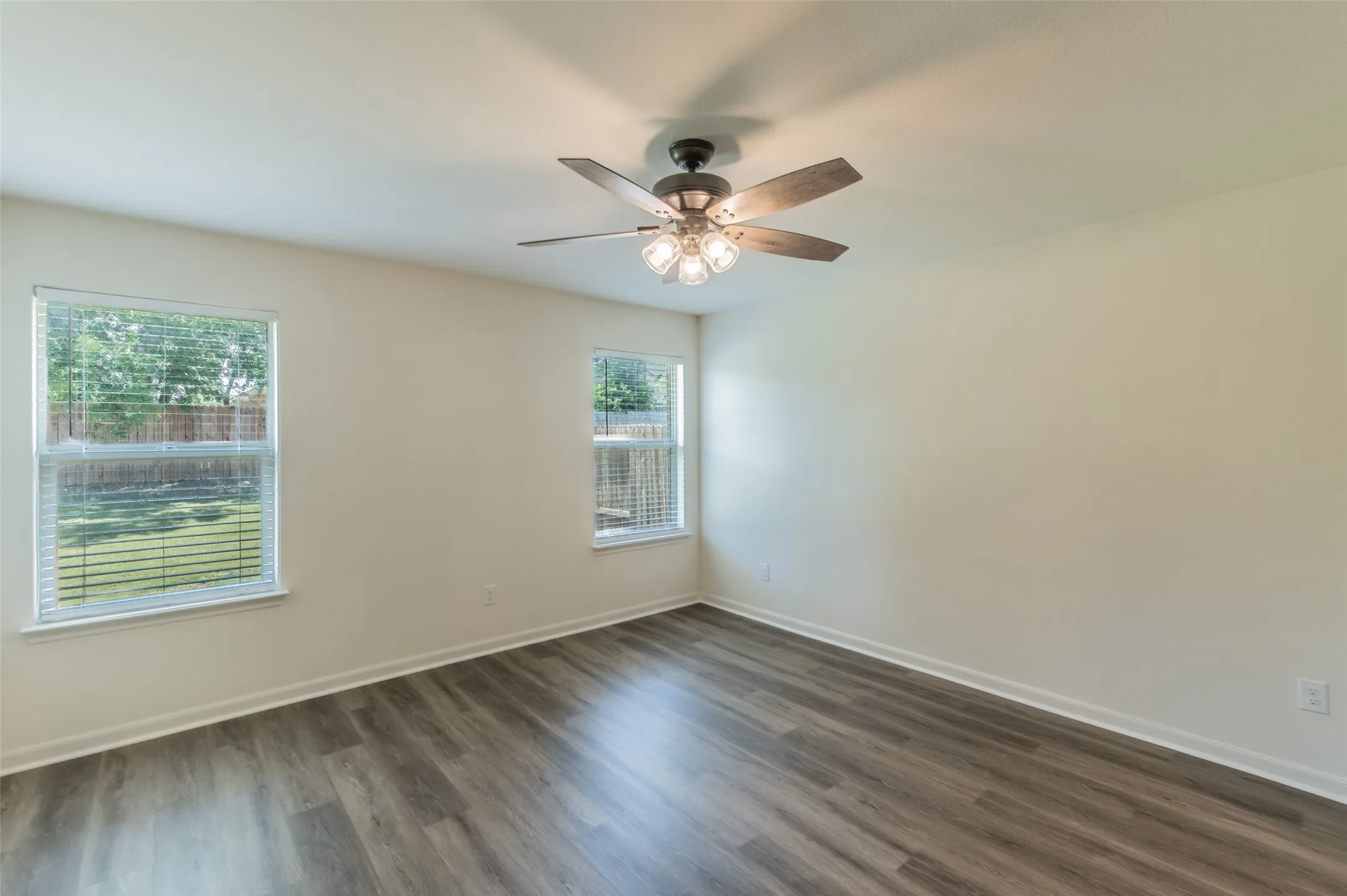 Spacious room featuring wood-finish flooring, two windows with blinds, a ceiling fan with integrated lighting, and neutral wall tones