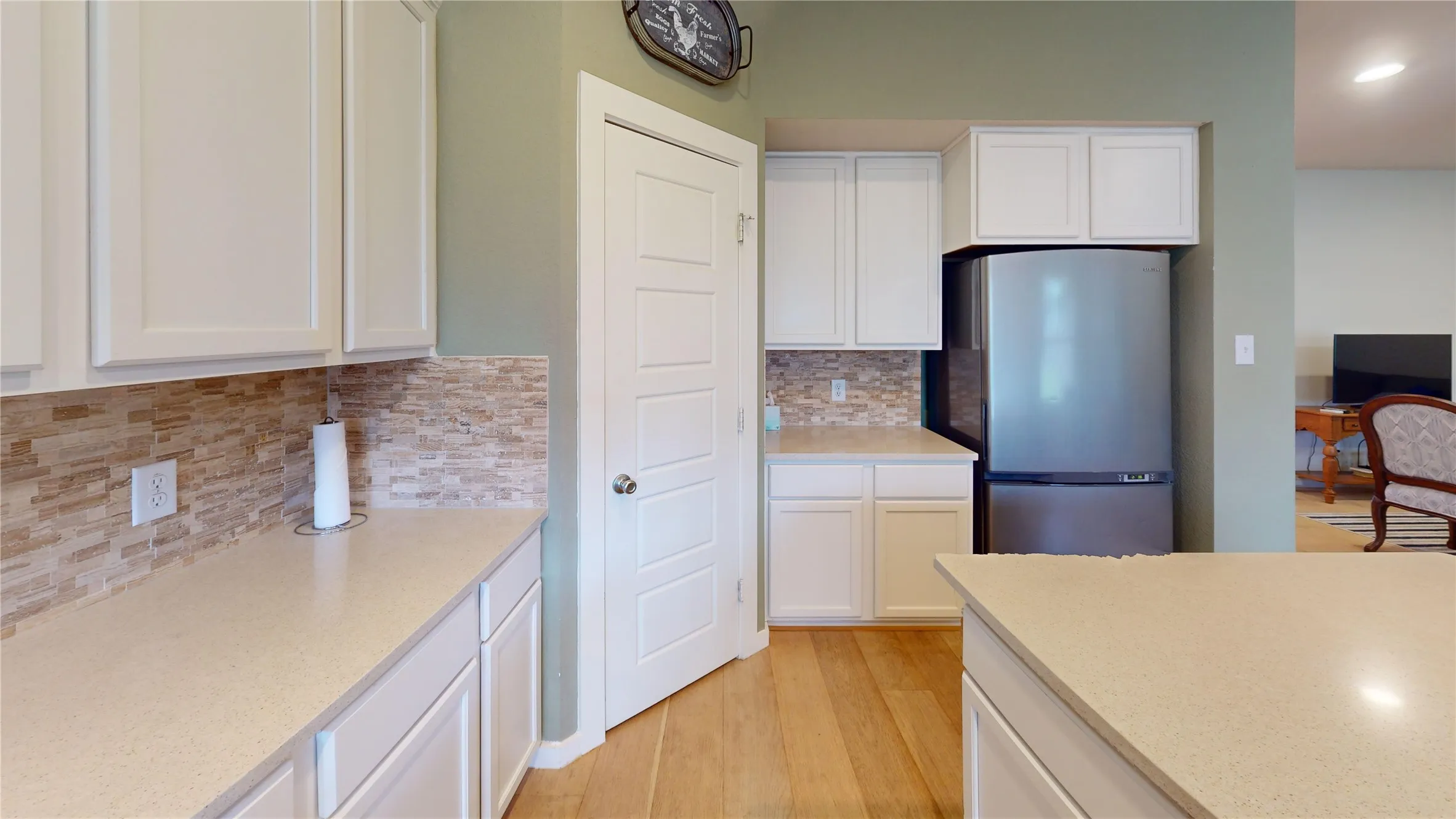 Kitchen featuring light-toned cabinetry, a textured tile backsplash, ample counter space, wood-finish flooring, and a stainless steel refrigerator