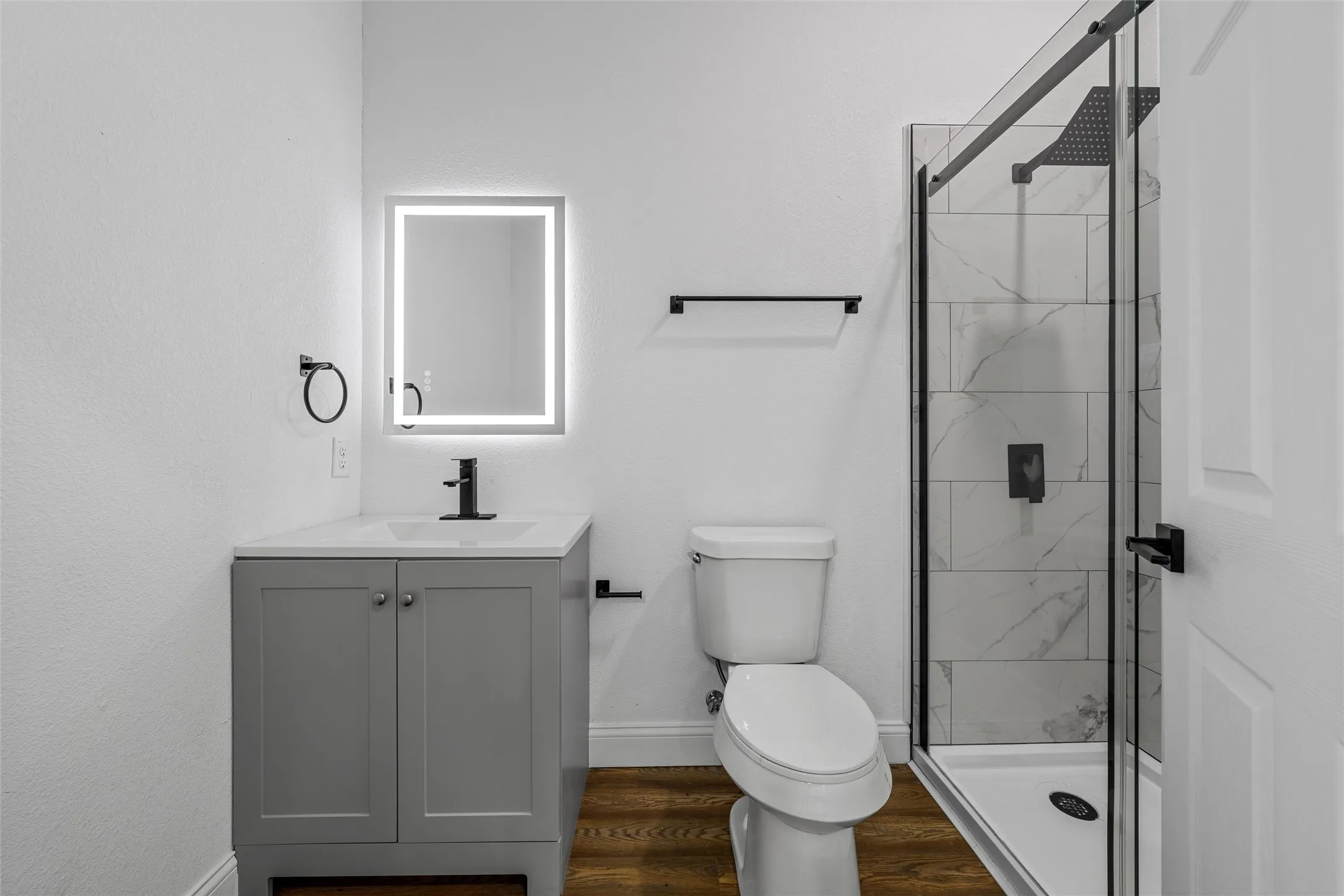Bathroom featuring a gray vanity with an integrated sink, an illuminated mirror, a glass-enclosed shower with tiled walls, and wood-finish flooring