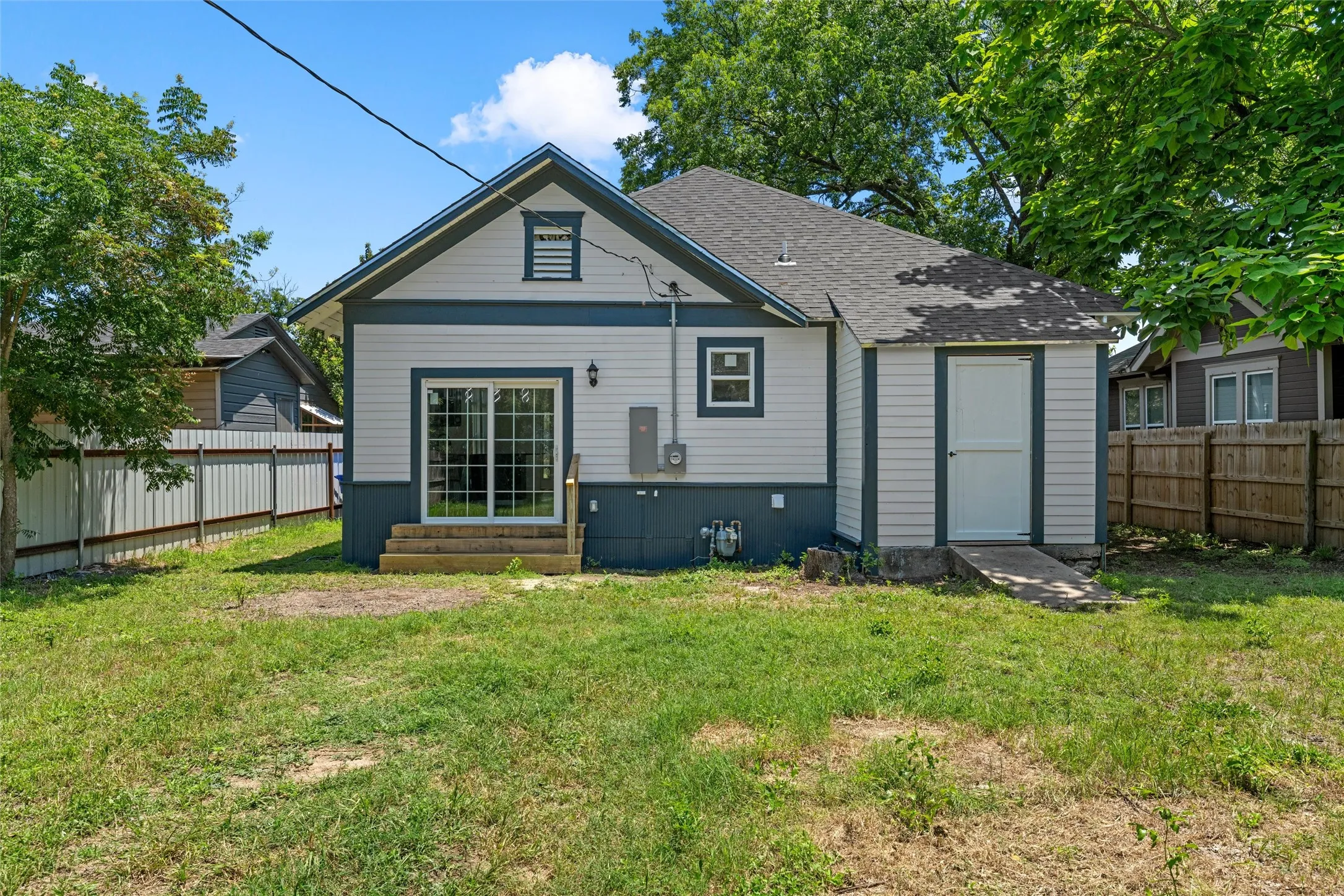 Rear exterior featuring white siding with dark trim, sliding glass doors, wooden steps, and an attached storage shed with a ramp