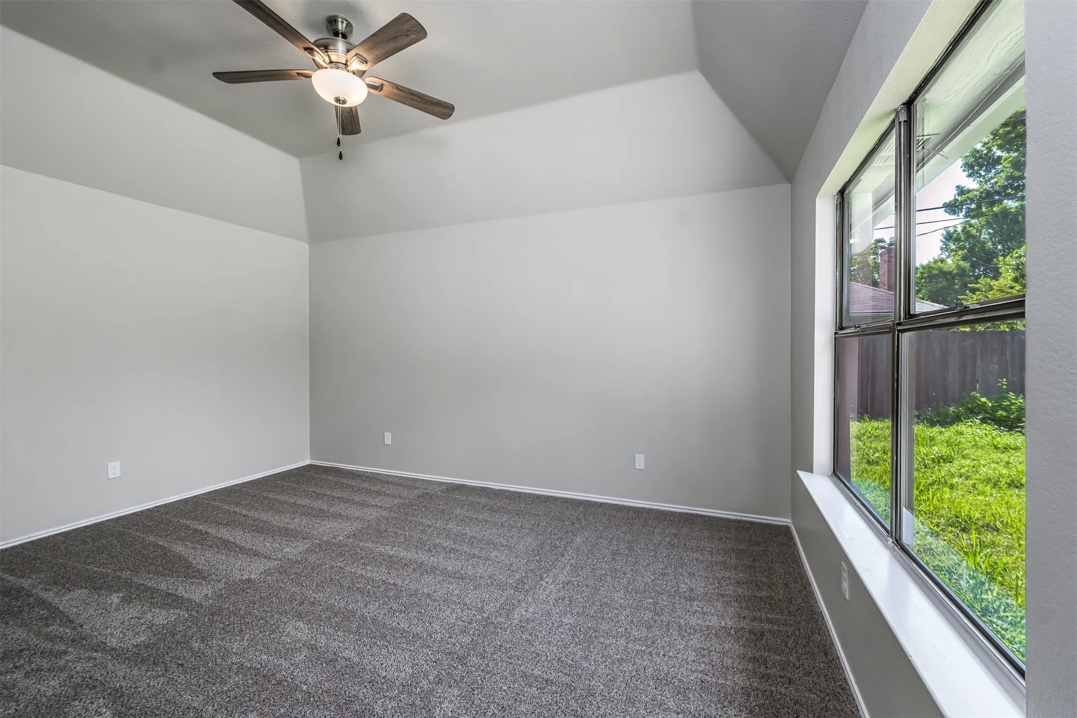 Spacious room featuring vaulted ceilings, a contemporary ceiling fan with integrated lighting, expansive window with a deep sill, neutral wall tones, and plush carpeting