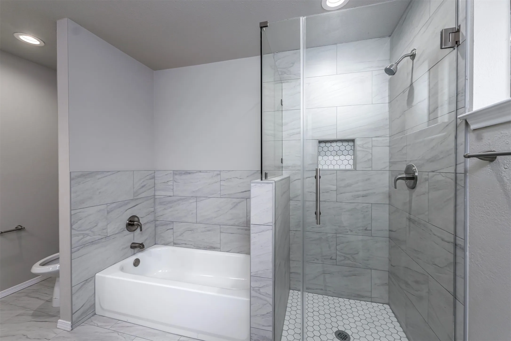 Bathroom featuring a white soaking tub, an adjacent glass-enclosed shower with a hexagon tile floor, a shower niche, and light-toned tile surrounds