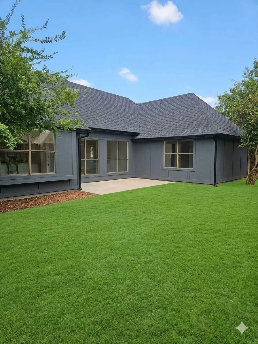 Expansive grassy area with a concrete patio, dark asphalt shingle roof, and multiple exterior windows