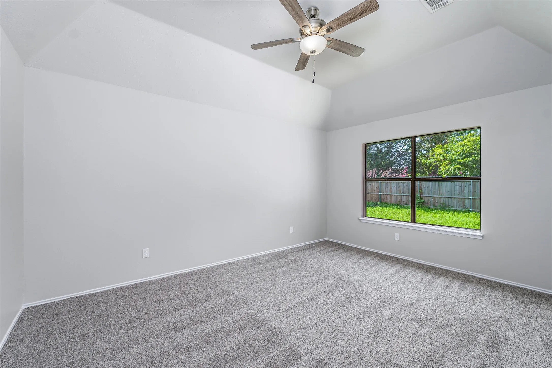 Spacious room featuring vaulted ceilings, a contemporary ceiling fan with integrated lighting, expansive window with exterior scenery, plush gray carpet, and clean white baseboards
