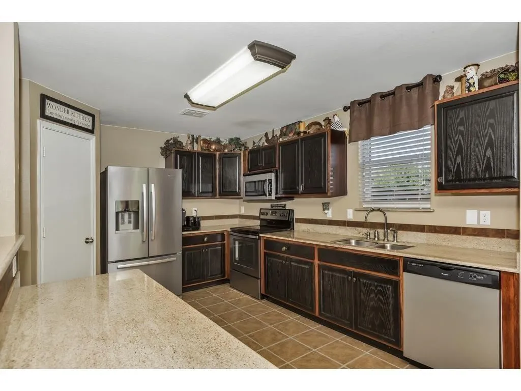 Kitchen featuring dark wood-finish cabinetry, light speckled countertops, a double basin stainless steel sink, and stainless steel appliances including a refrigerator and dishwasher