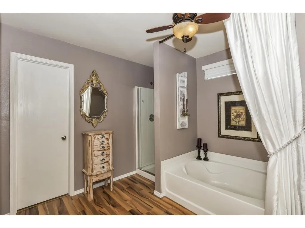 Bathroom featuring a built-in soaking tub, glass-enclosed shower, wood-finish flooring, ceiling fan with light fixture, and a window with blinds