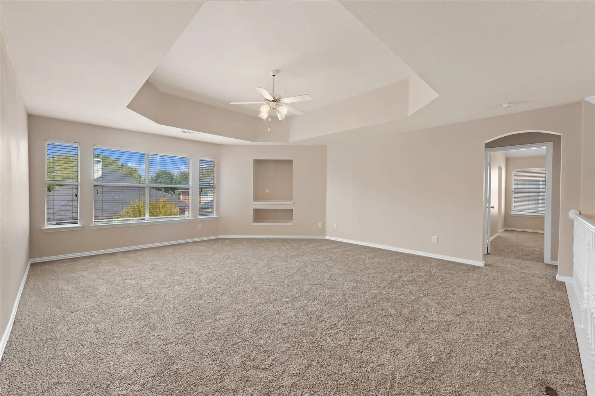 Spacious room featuring neutral carpeting, a tray ceiling with a ceiling fan, multiple windows with blinds, built-in wall niches, and an arched doorway