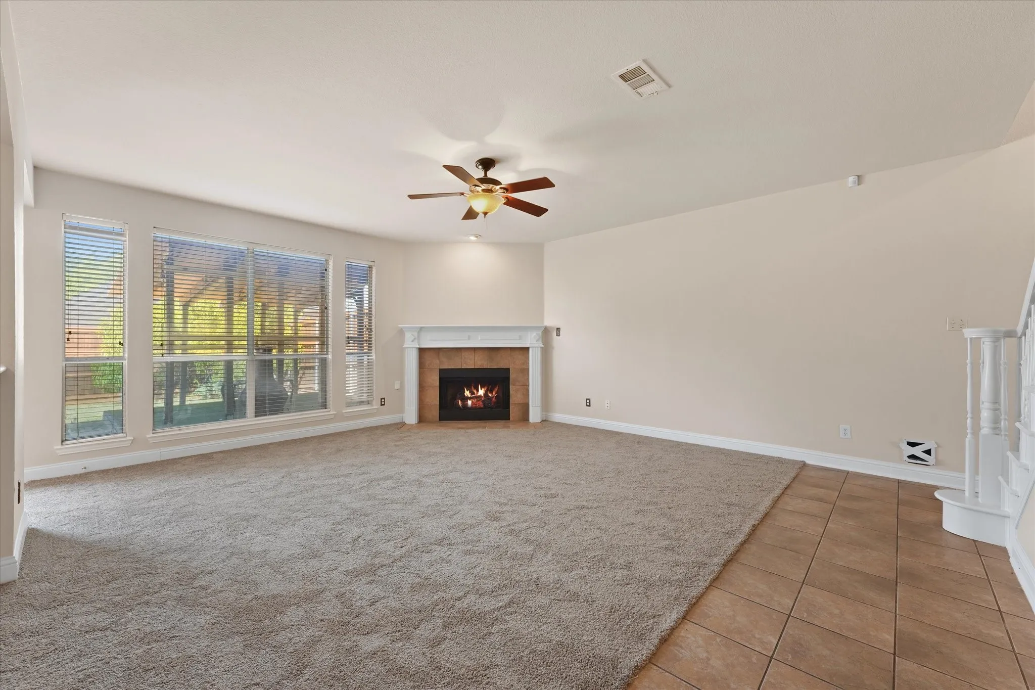 Spacious living area featuring a fireplace with a white mantel, multiple large windows, a ceiling fan, and a combination of carpet and tile flooring