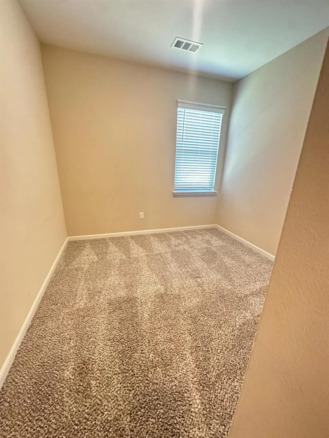 Carpeted room featuring neutral wall paint, a single window with blinds, white baseboards, and a ceiling-mounted vent