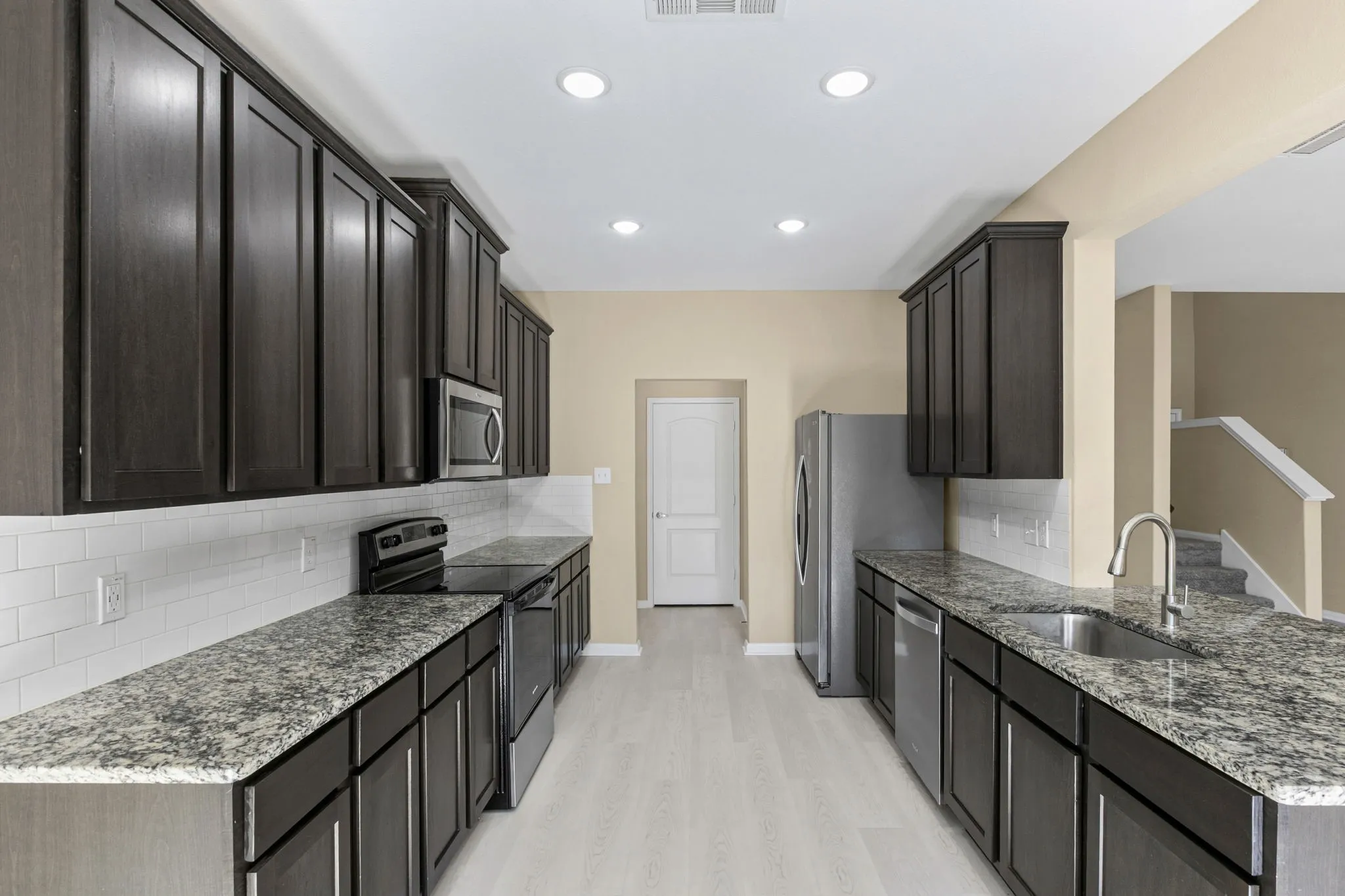 Galley kitchen featuring dark wood-finish cabinetry, granite-style countertops, stainless steel appliances, and white subway tile backsplash
