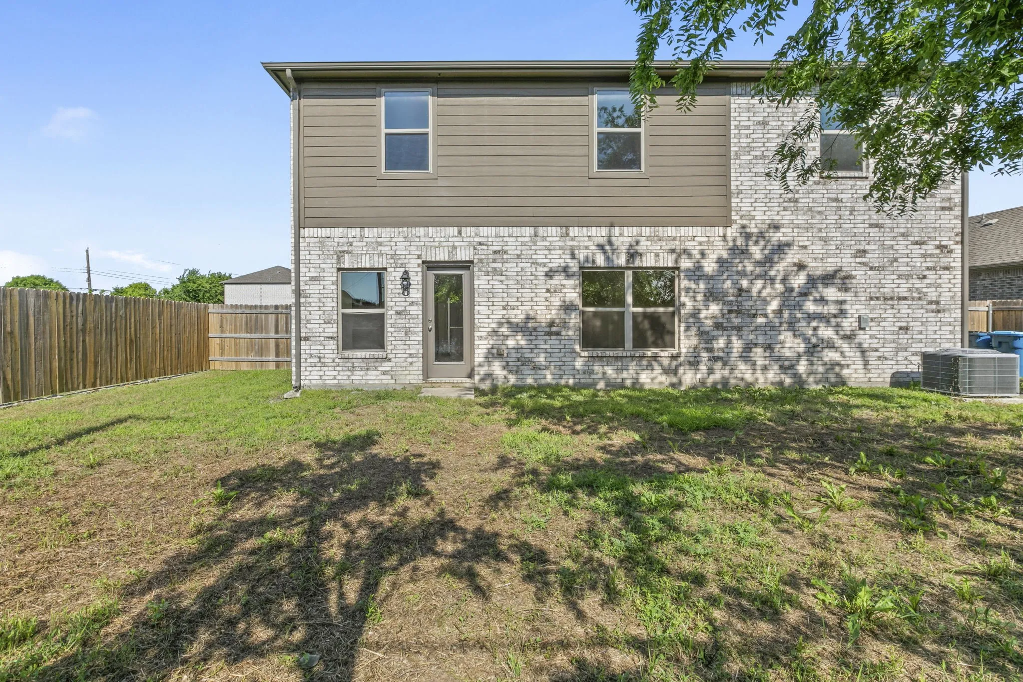 Rear exterior featuring a mix of light-toned brick and horizontal siding, multiple windows, a rear entry door with an exterior light fixture, and a wood privacy fence