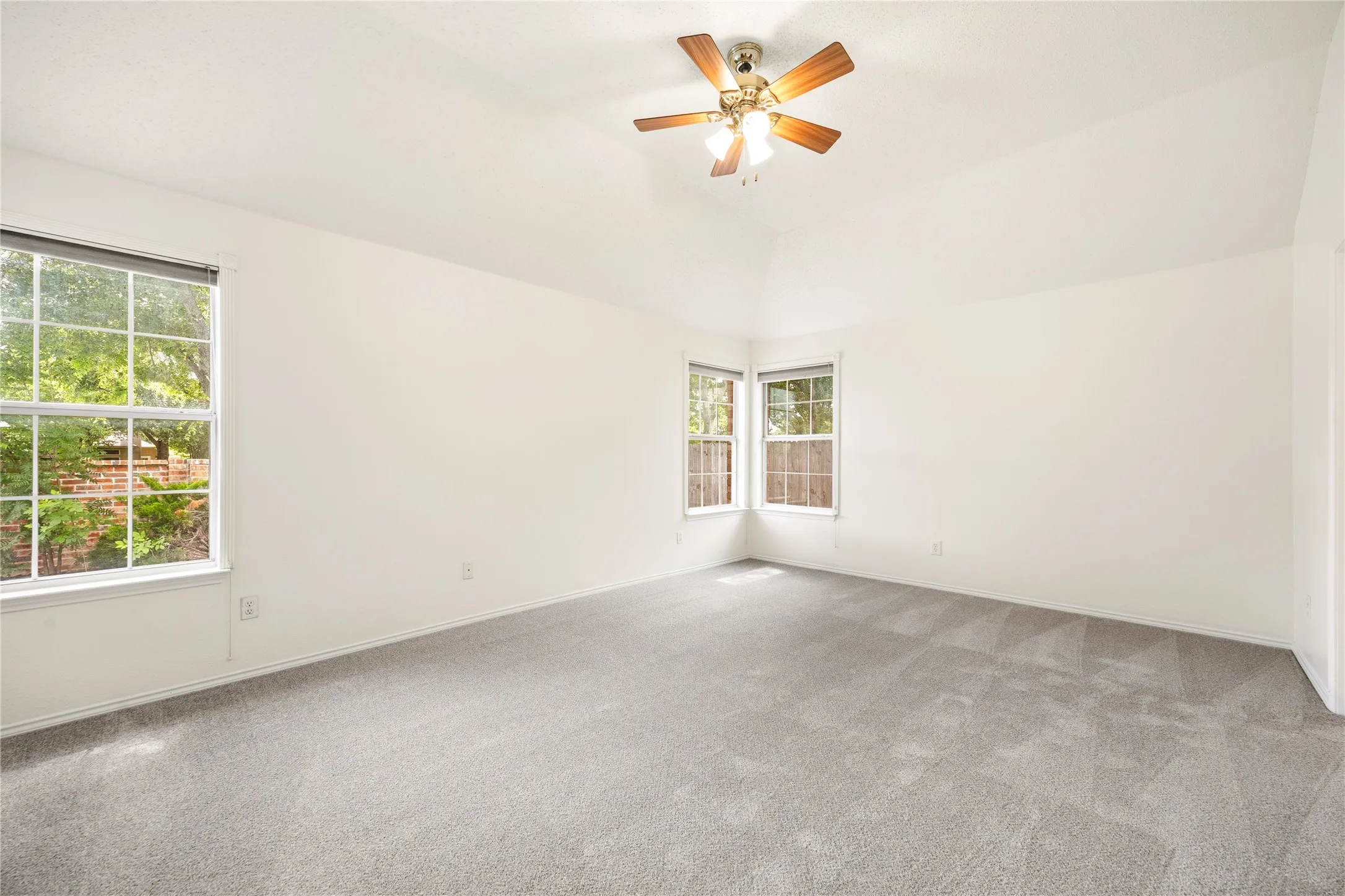 Spacious interior featuring a vaulted ceiling, gray carpet flooring, multiple windows providing natural light, and a ceiling fan with light fixture