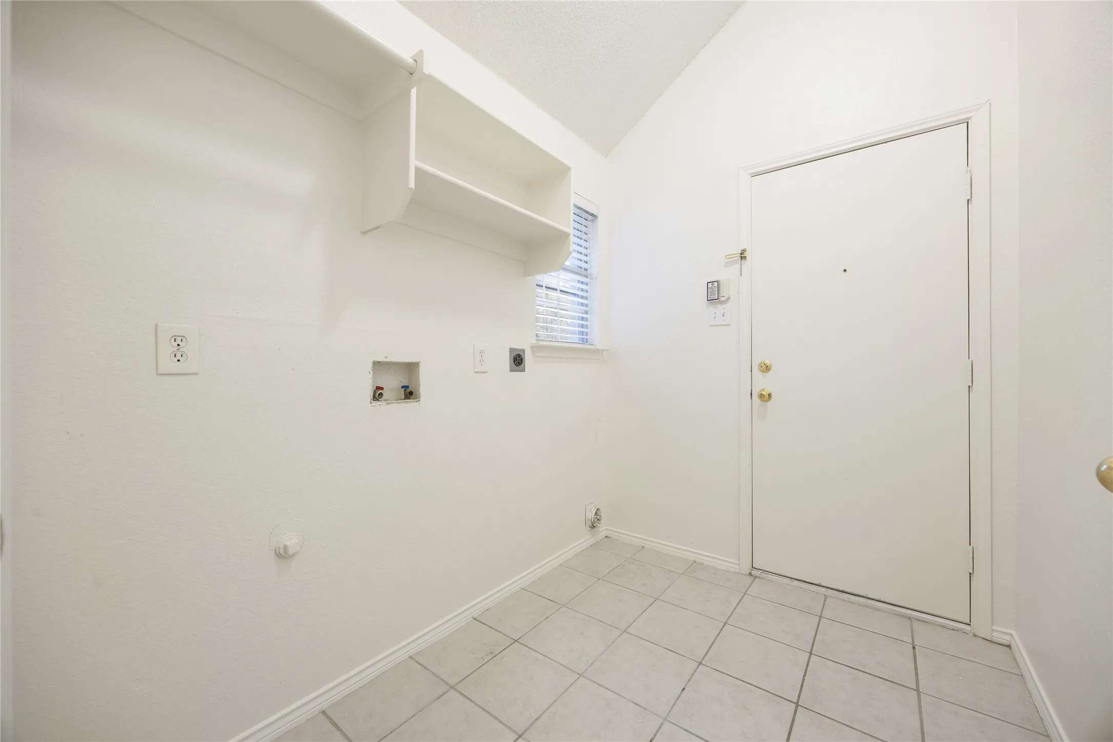 Utility space featuring tile flooring, built-in shelving, a window with blinds, and a solid entry door with brass hardware