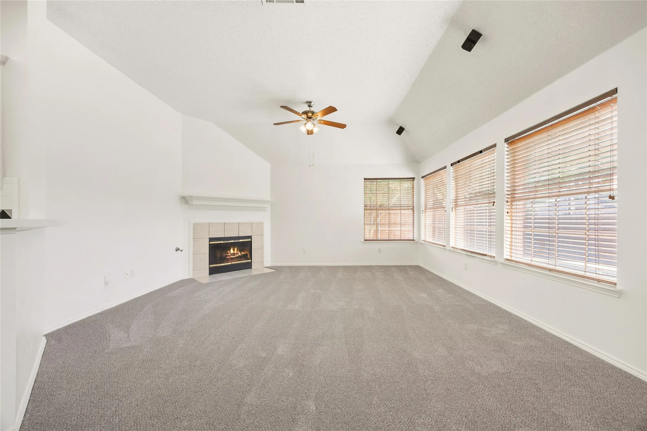 Spacious living area featuring a vaulted ceiling with a ceiling fan, a tiled fireplace with a mantel, and multiple windows with blinds