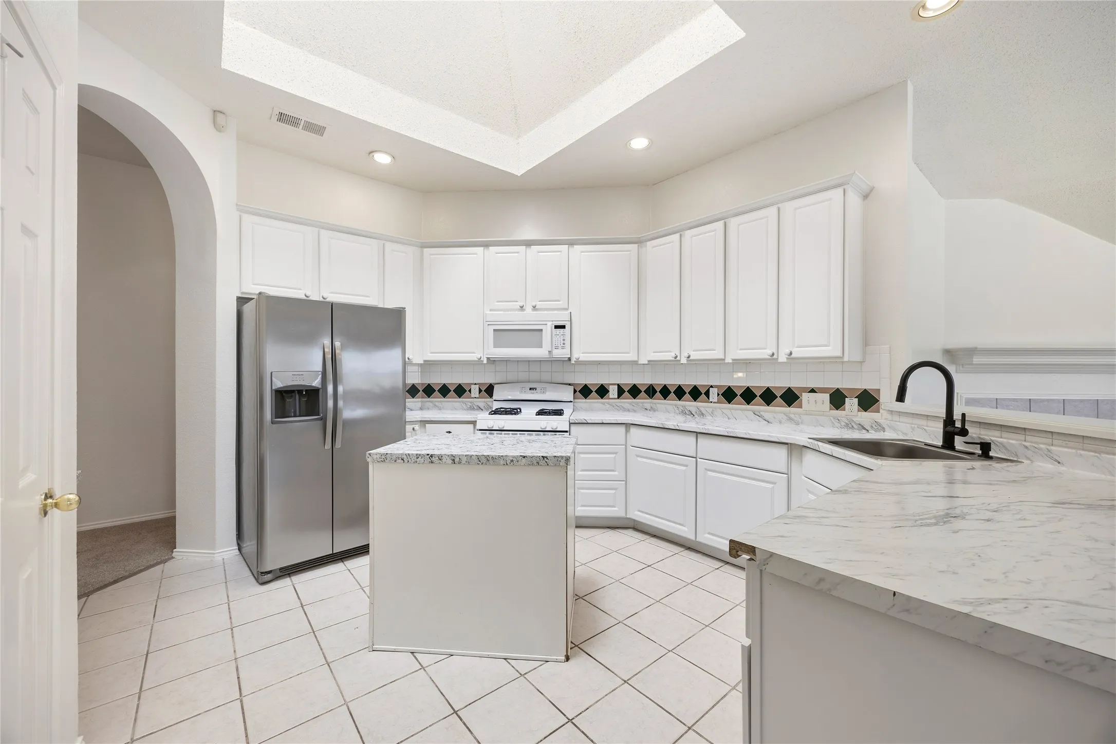 Kitchen featuring white cabinetry, light-toned countertops, tile flooring, stainless steel refrigerator, and an arched entryway