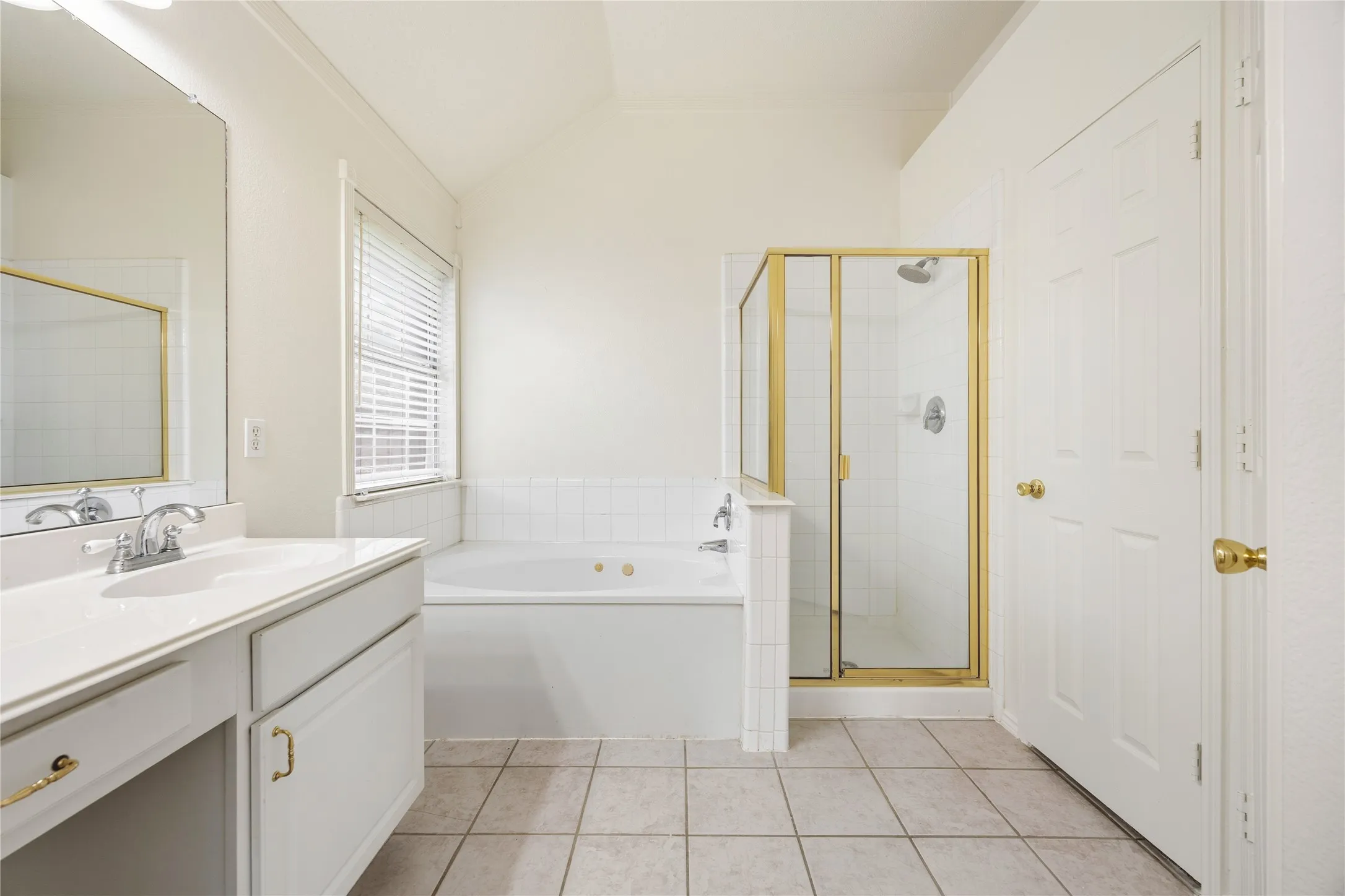 Spacious bathroom featuring a white vanity with integrated sink, a large soaking tub with tile surround, a separate shower with a gold-framed glass enclosure, and neutral tile flooring