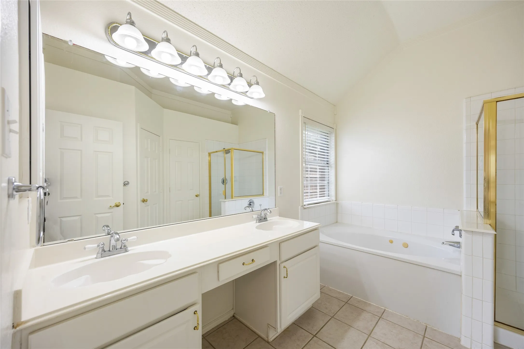 Bathroom featuring a double vanity with integrated sinks, a built-in jetted tub, a separate shower with a gold-toned frame, and neutral tile flooring