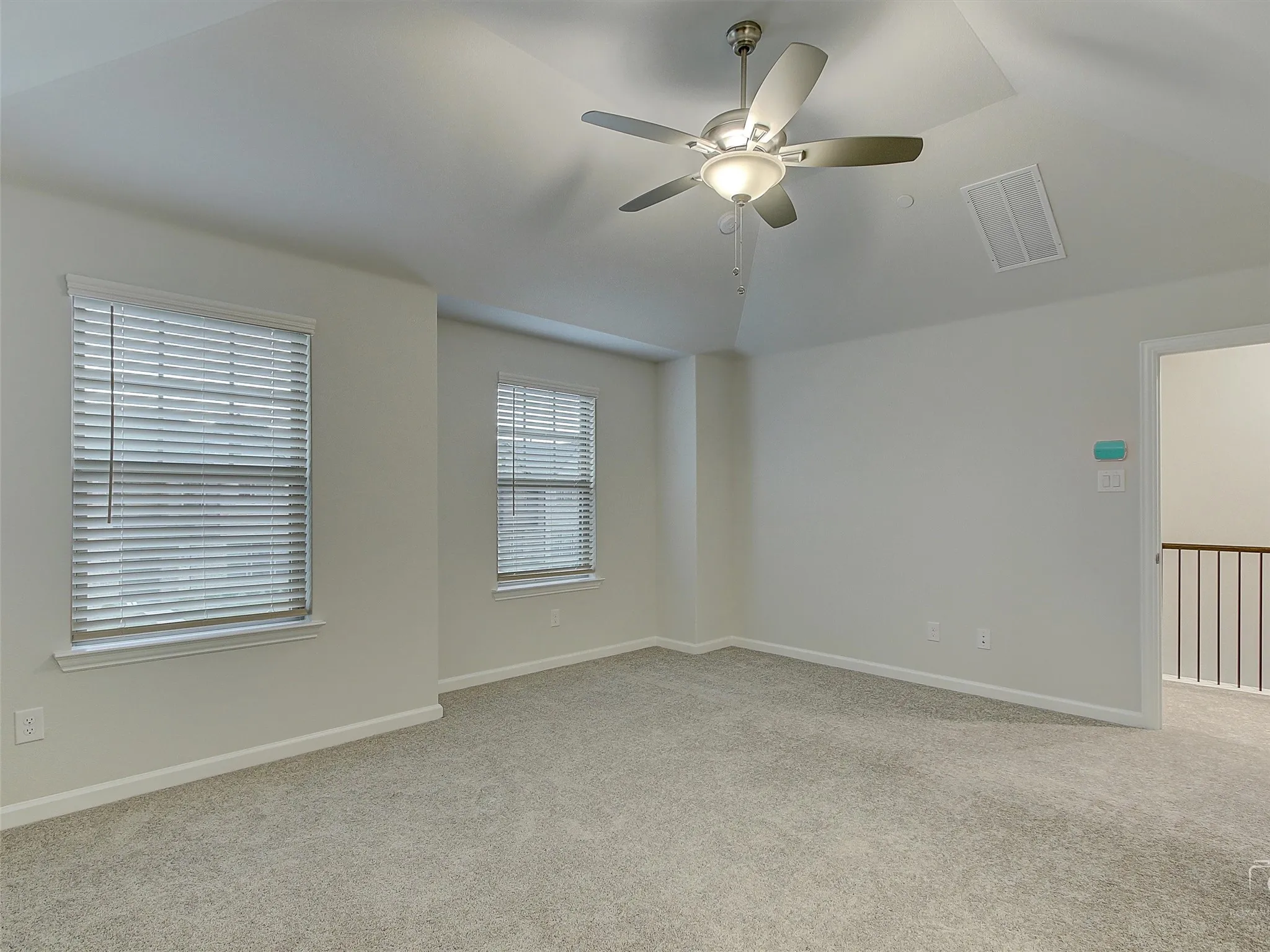 Carpeted room featuring a ceiling fan with integrated lighting, two windows with horizontal blinds, a vaulted ceiling, and a doorway opening to a landing with a metal spindle railing
