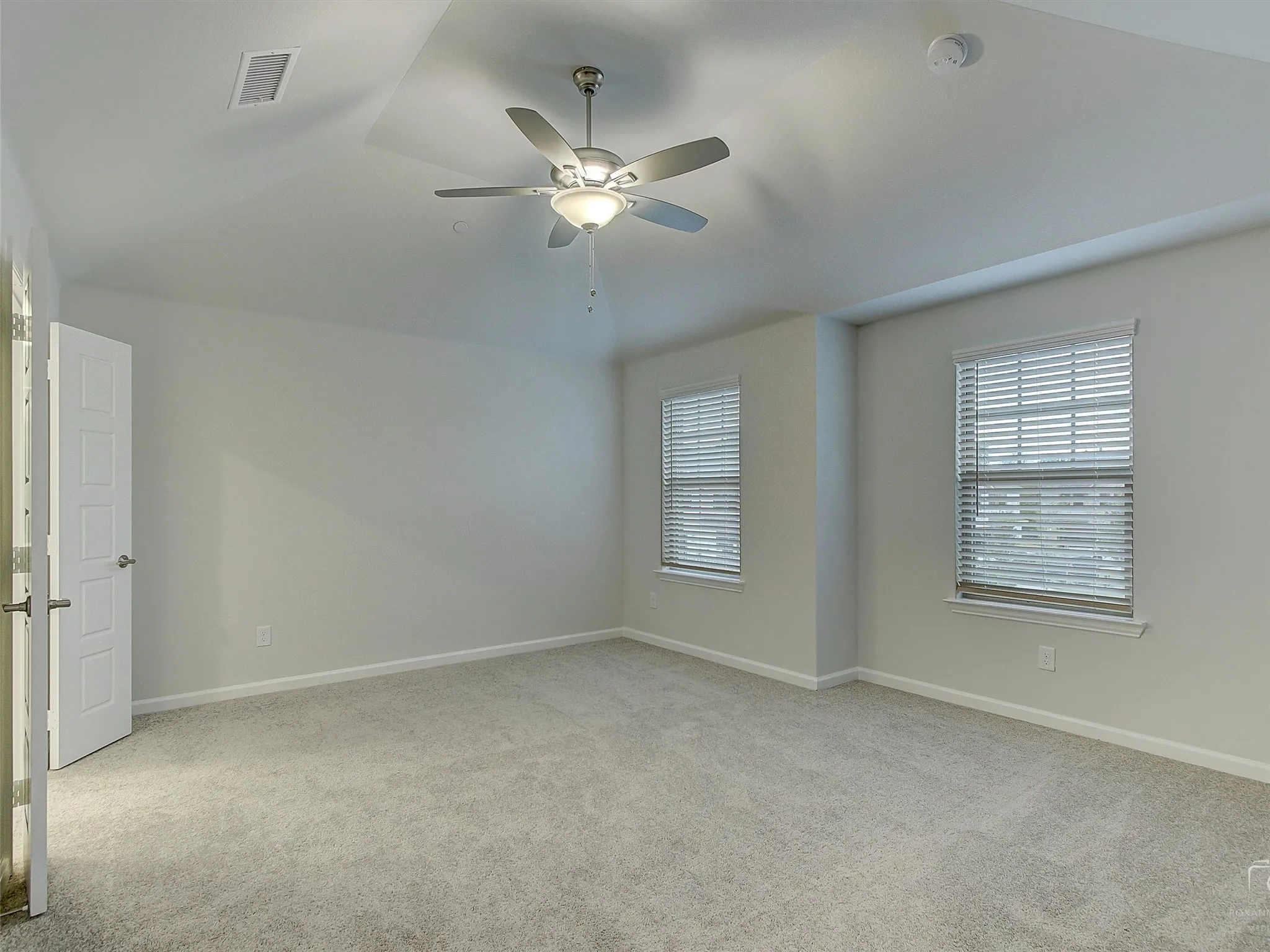 Expansive room featuring a tray ceiling, ceiling fan with integrated lighting, two windows with horizontal blinds, and neutral carpeting