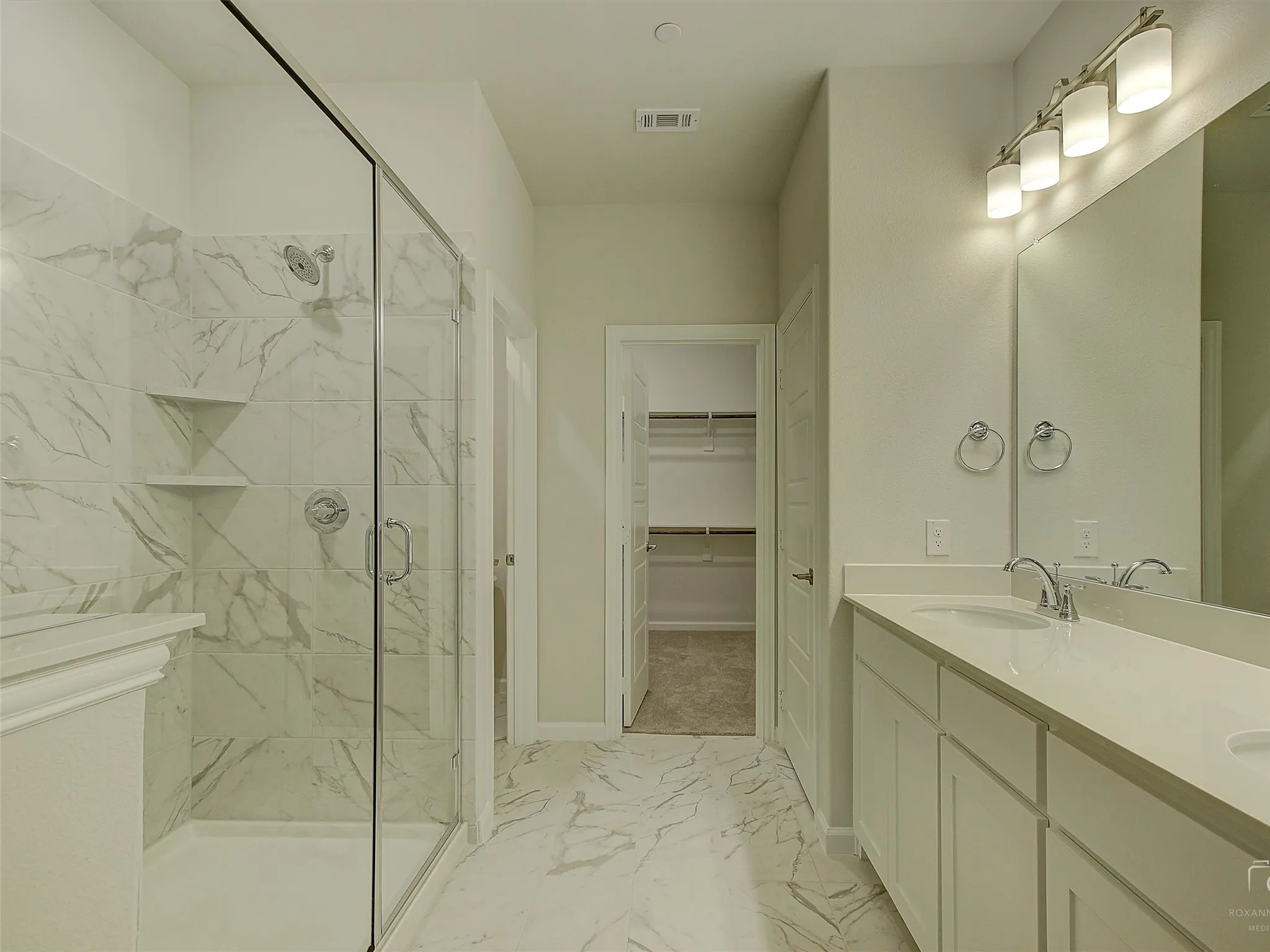 Spacious bathroom featuring a glass-enclosed shower with tiled walls and built-in shelving, a dual vanity with a light-colored countertop and undermount sinks, and a walk-in closet with built-in shelving