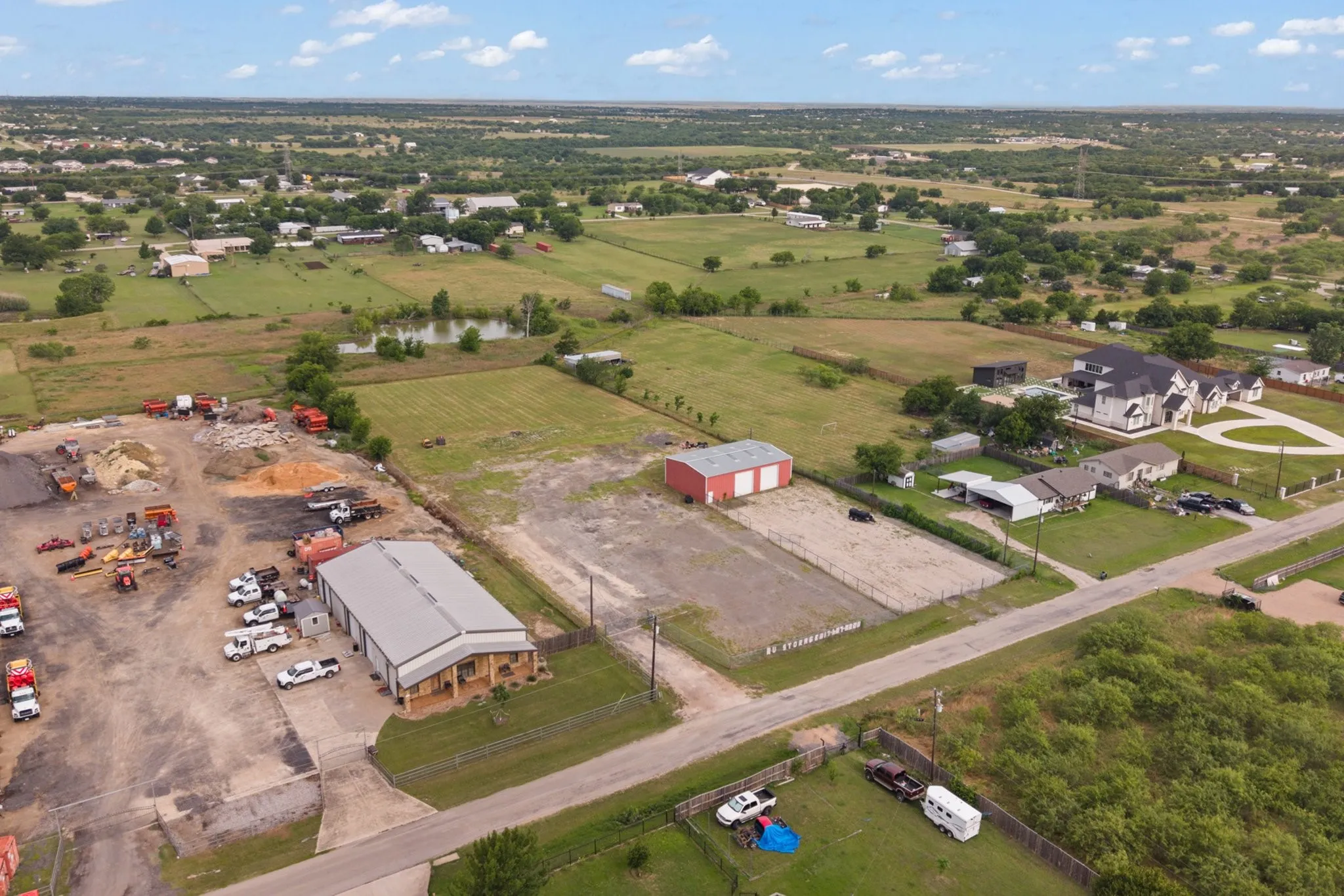 Expansive rural property featuring a large commercial-style building with a metal roof and covered porch, a separate red barn structure, undeveloped land parcels, and a visible pond