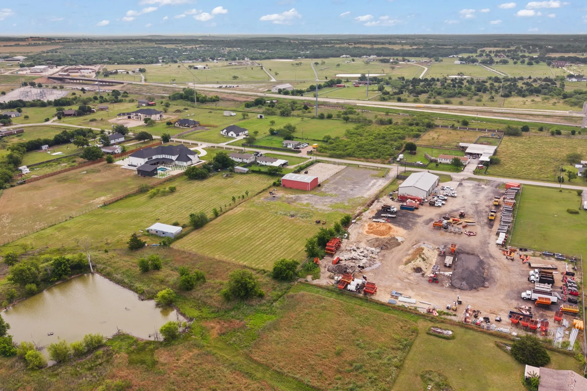 Expansive aerial view showcasing a large commercial lot with multiple structures and equipment, a nearby pond, and surrounding open land