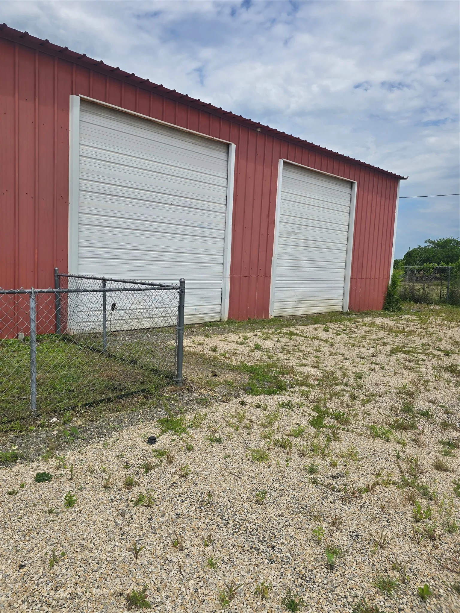 Red metal-sided building featuring two large white roll-up doors, a sloped metal roofline, and a partial chain-link fence