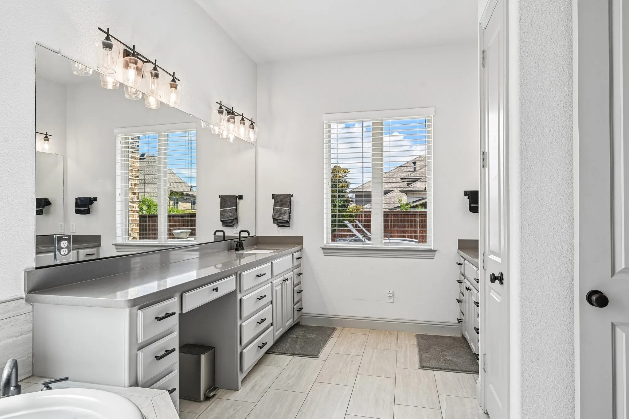 Dual vanities with undermount sinks and matte black fixtures, extensive cabinetry with matte black hardware, large mirror with sconce lighting, white tiled flooring, and a large window with privacy blinds